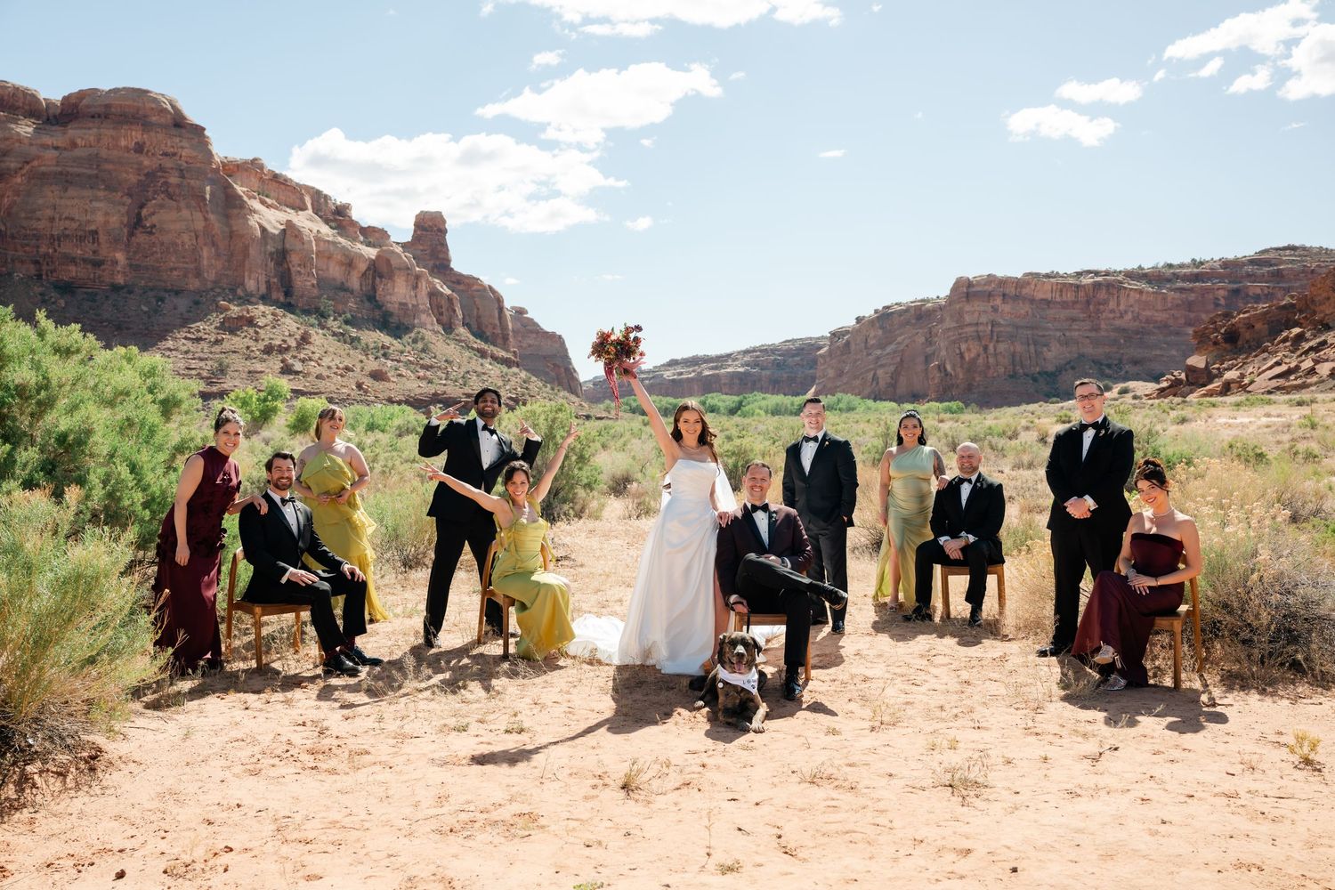 Wedding party poses in stunning desert canyon landscape with red rock formations under a bright blue sky.