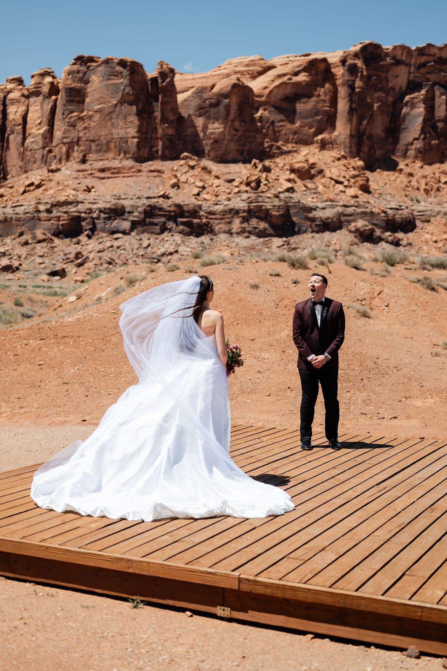 Bride in flowing white gown and veil stands on wooden platform with groom in desert landscape with red rock during emotional first look.