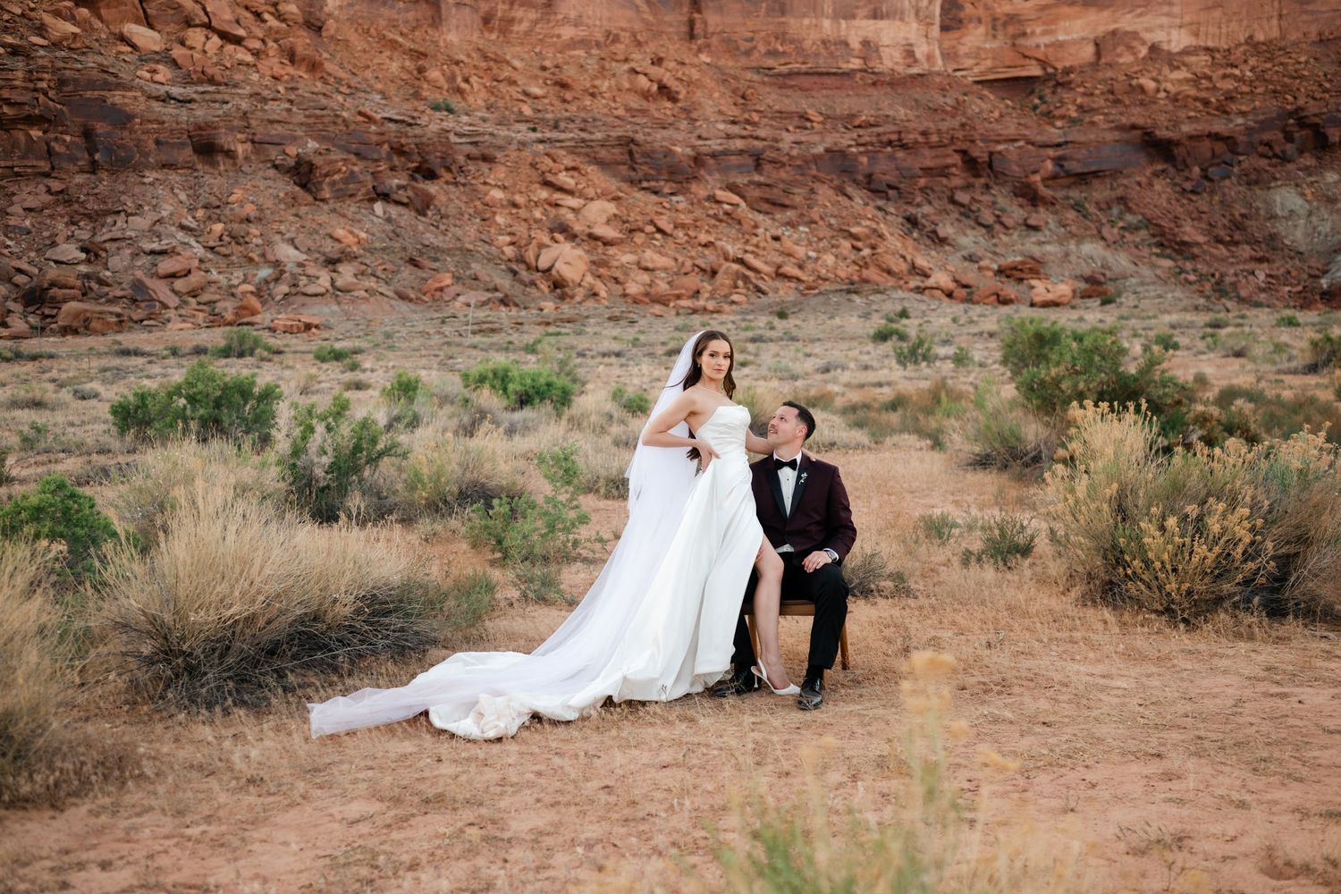 Bride in white gown and groom in tuxedo pose romantically in a desert landscape with red rock formations.