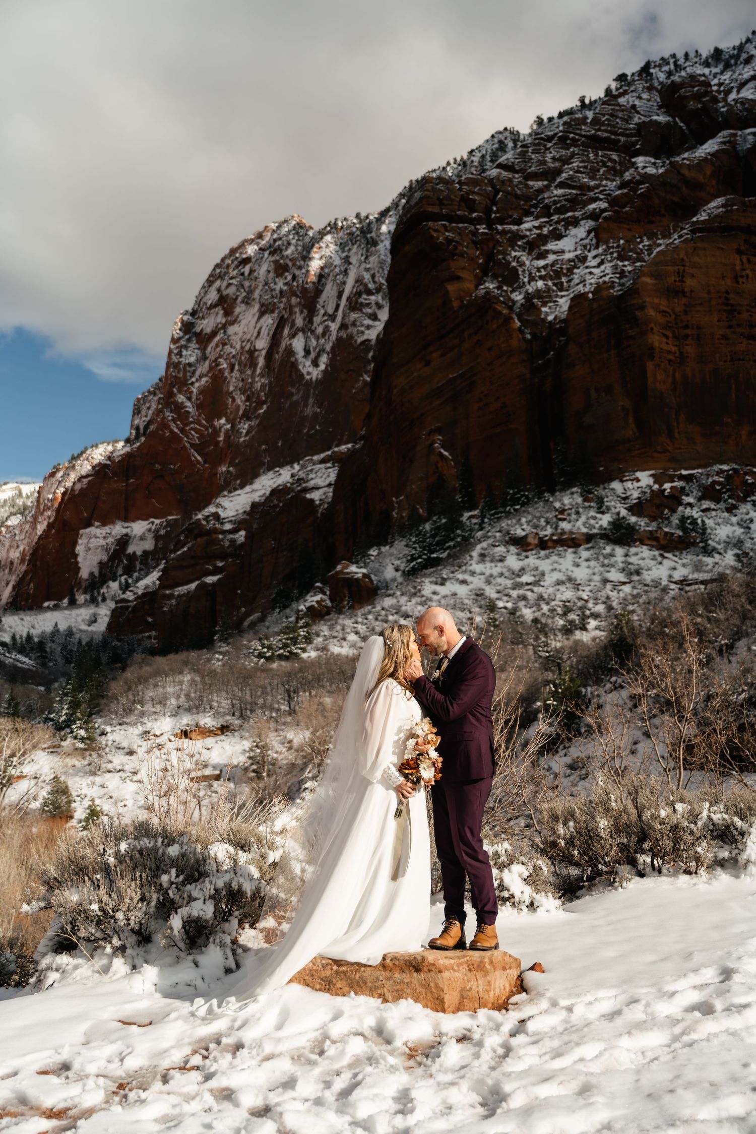 A winter wedding photoshoot against snow-covered red rock cliffs with dramatic mountain scenery.