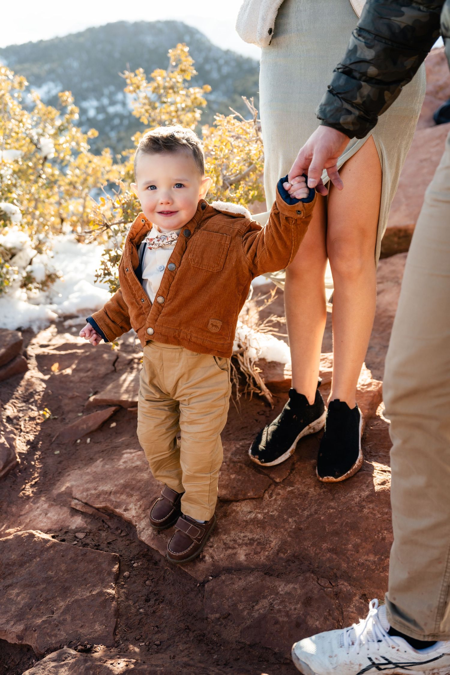 A small child in brown overalls walks along a rocky trail with adults in the background.