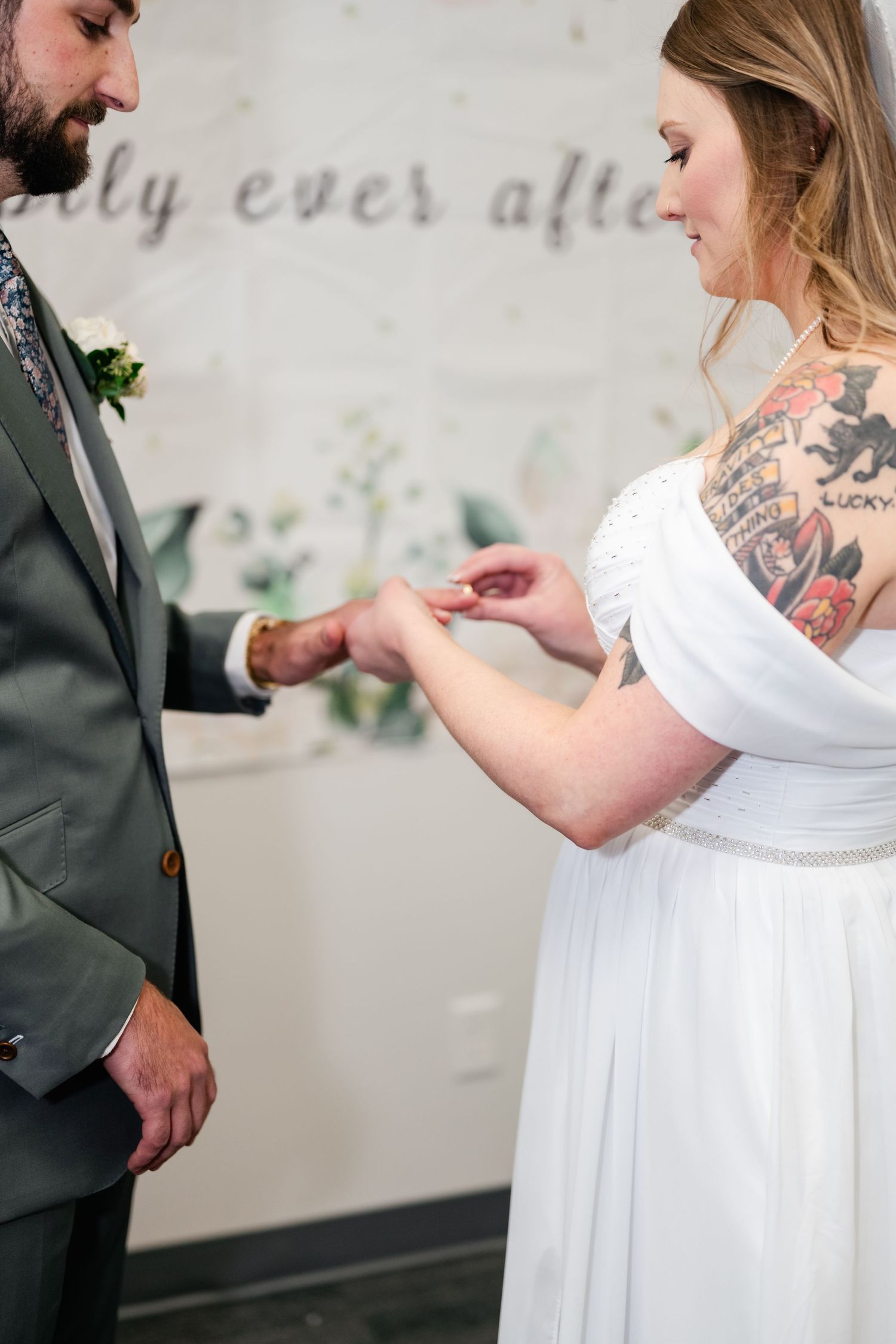 A wedding detail shot showing a couple exchanging rings in front of a wall with elegant script lettering.
