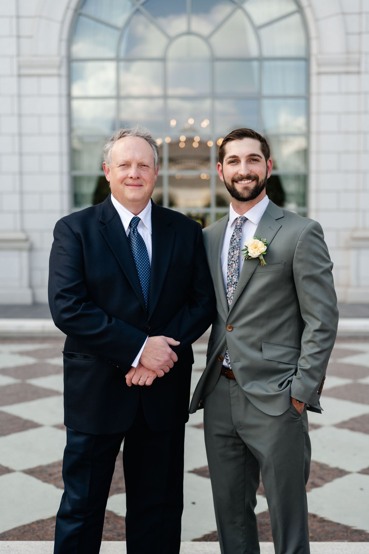 Two people in formal attire stand together in front of an elegant arched window with a chandelier visible.