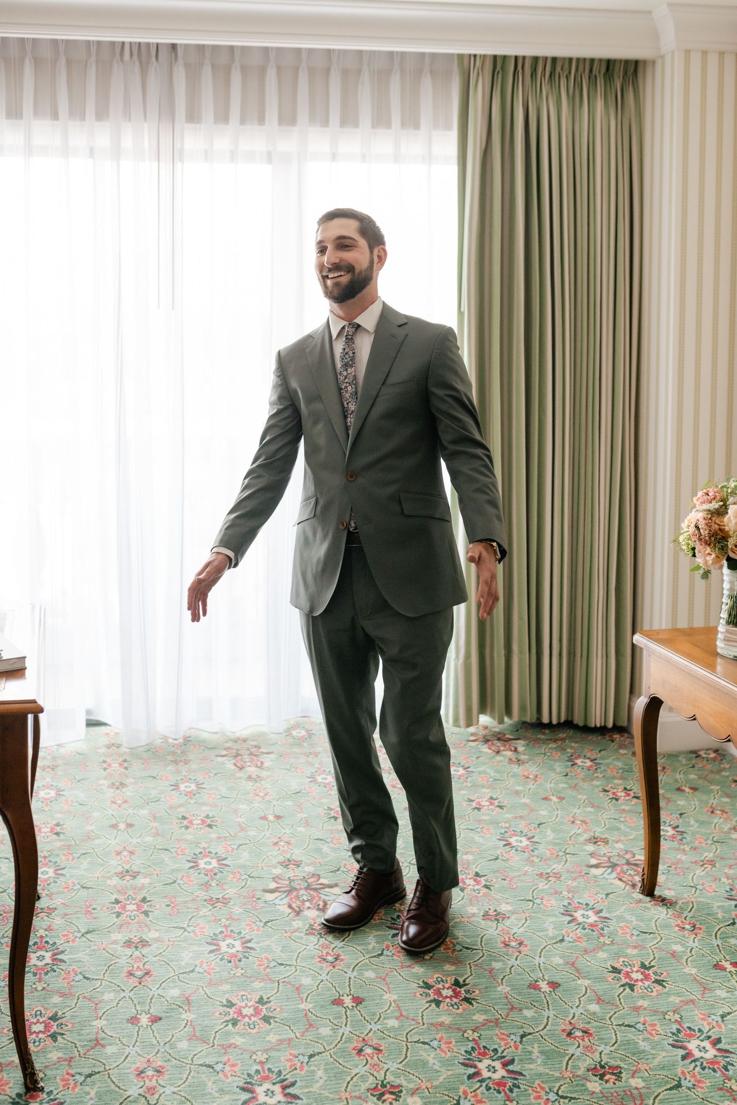 Person in a grey suit walking on floral patterned carpet in a bright hotel room.