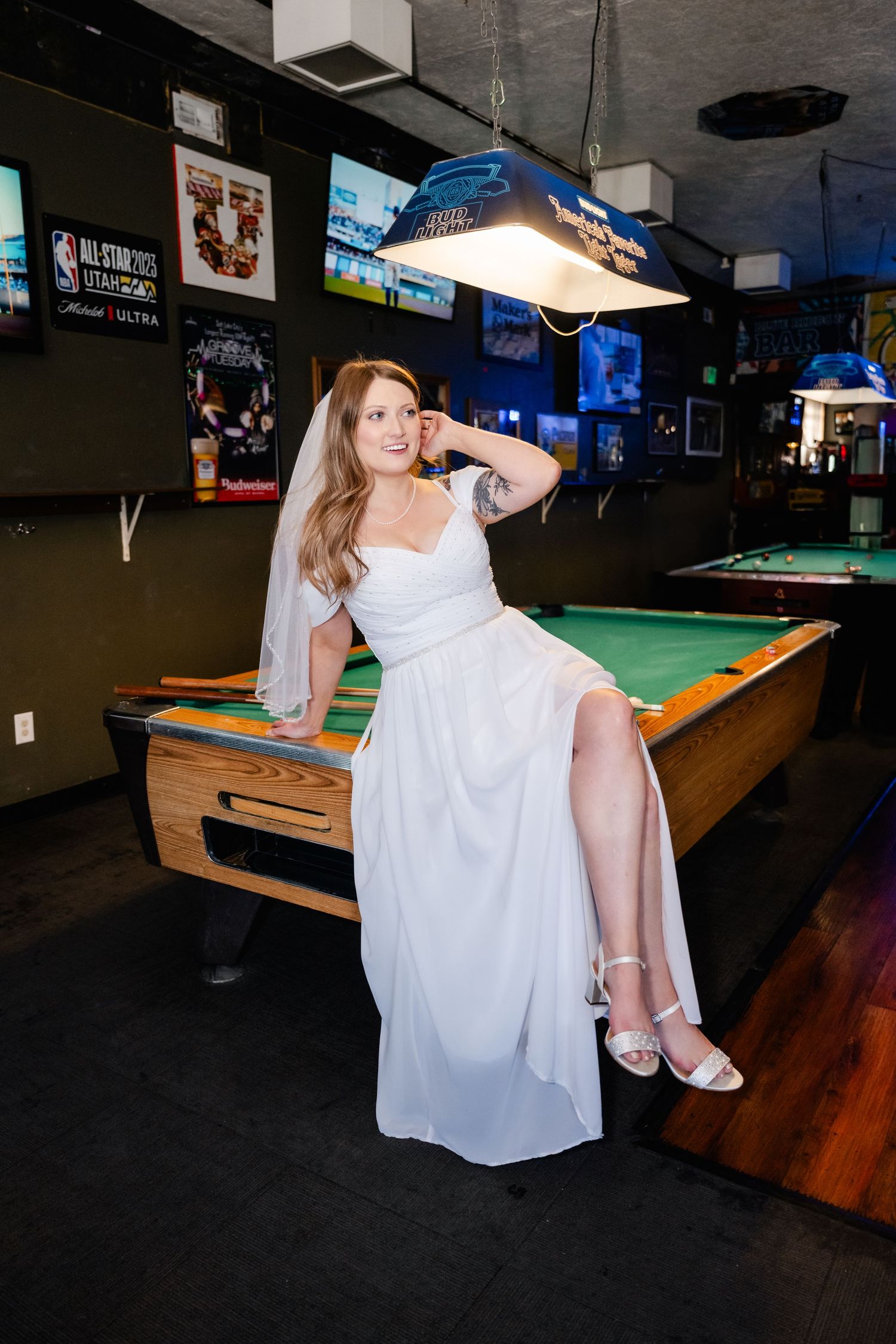 A bride in a white dress poses playfully on a pool table during a wedding reception.