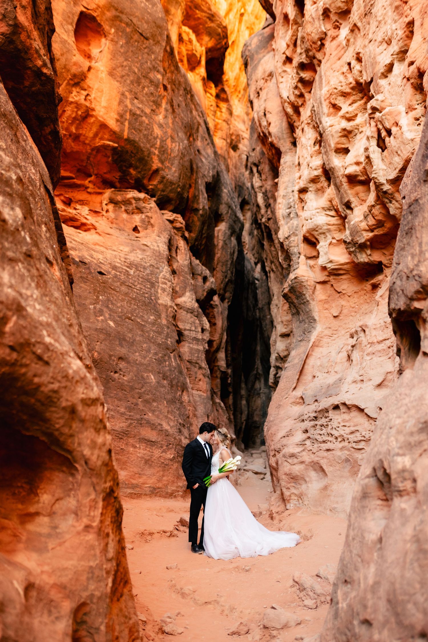 A wedding couple poses for romantic photos inside a dramatic red rock canyon cave formation. Snow Canyon Wedding Photographer.