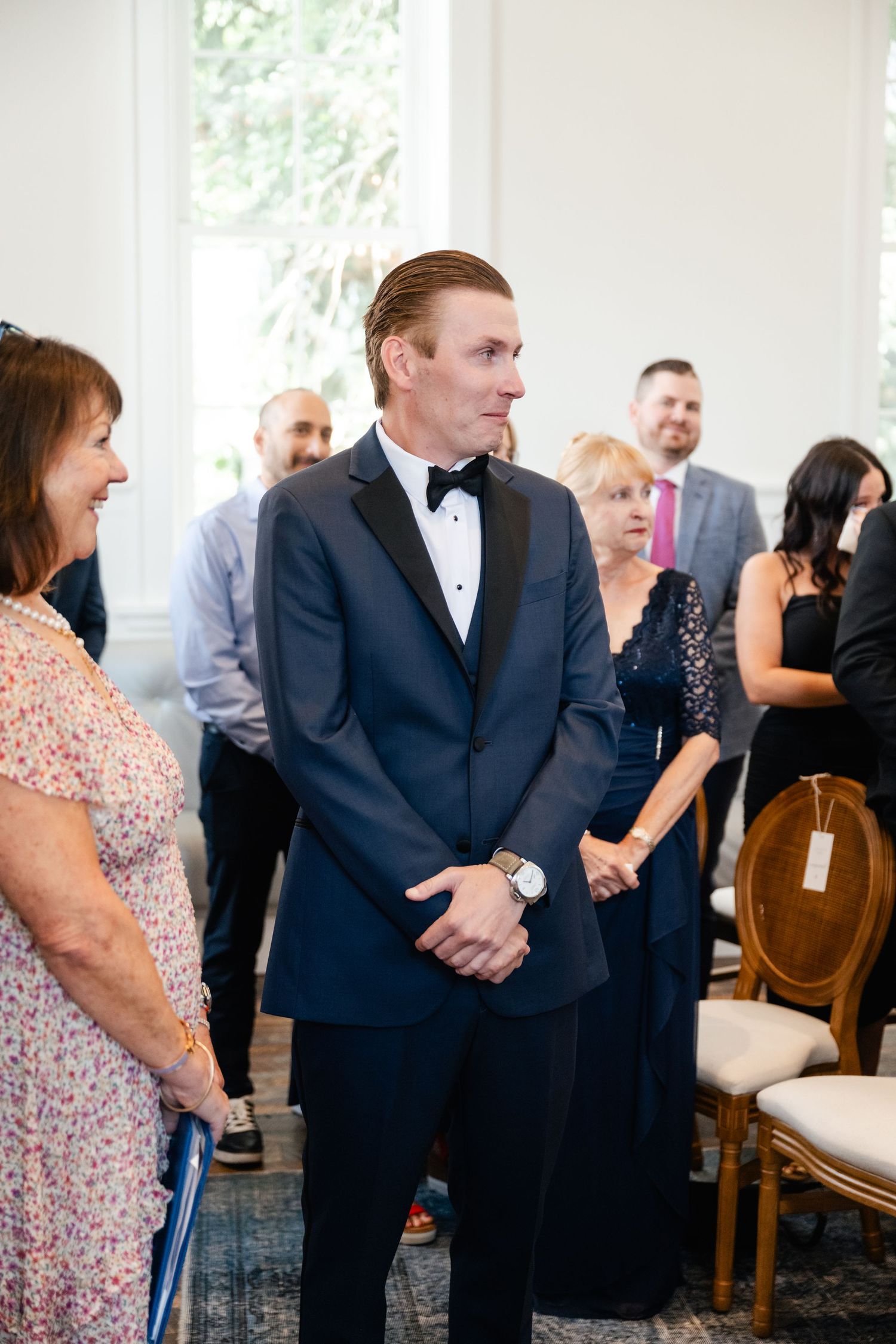 Guests in formal attire including dark suits and floral dresses gather at an elegant indoor event space.