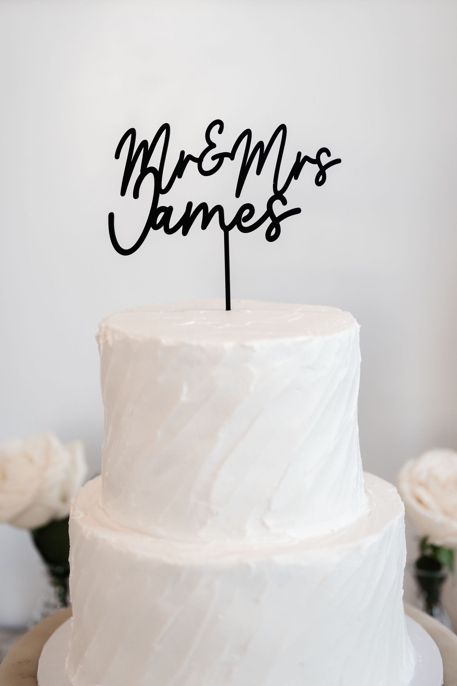 White tiered wedding cake with black calligraphy topper reading 'Mr & Mrs' against light background.
