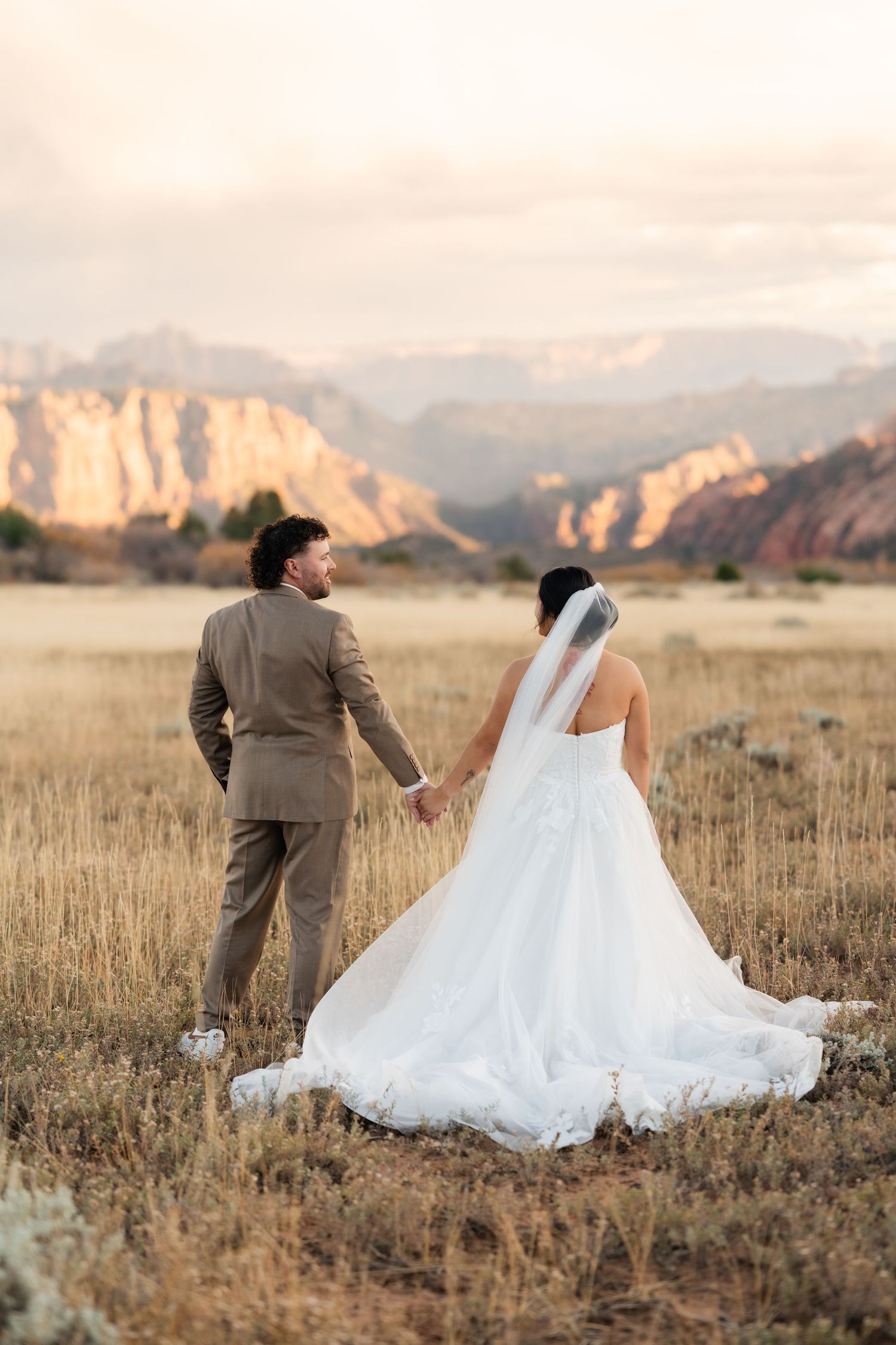 A wedding couple poses in a golden field with dramatic mountain backdrop during sunset.