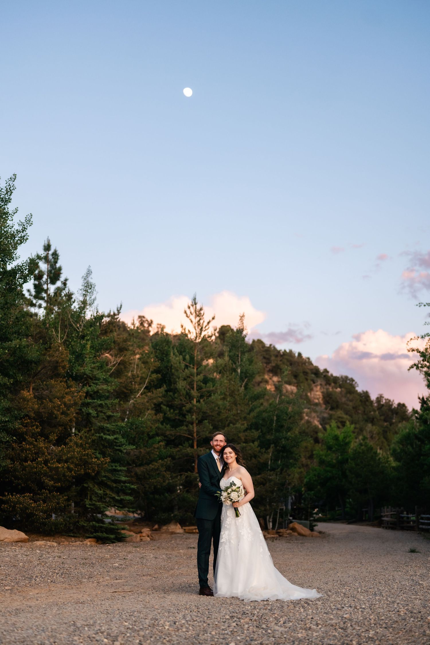 A couple in wedding attire poses against a scenic mountain backdrop at sunset with pine trees and desert landscape.