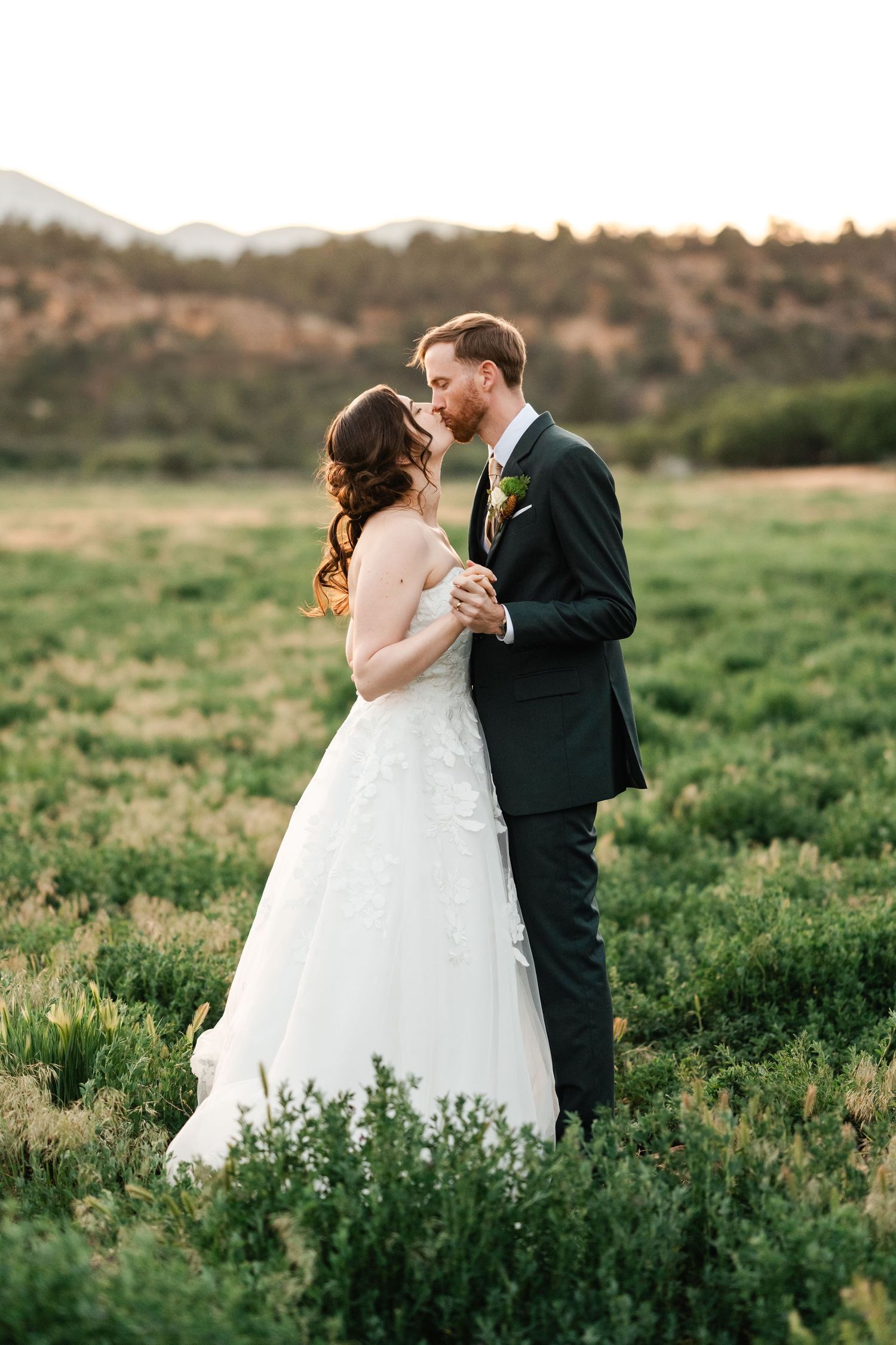 Romantic wedding couple embraces in a green meadow at sunset.