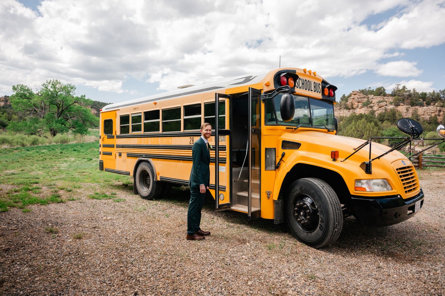 Line of yellow school buses parked on dirt road with mountains in background.
