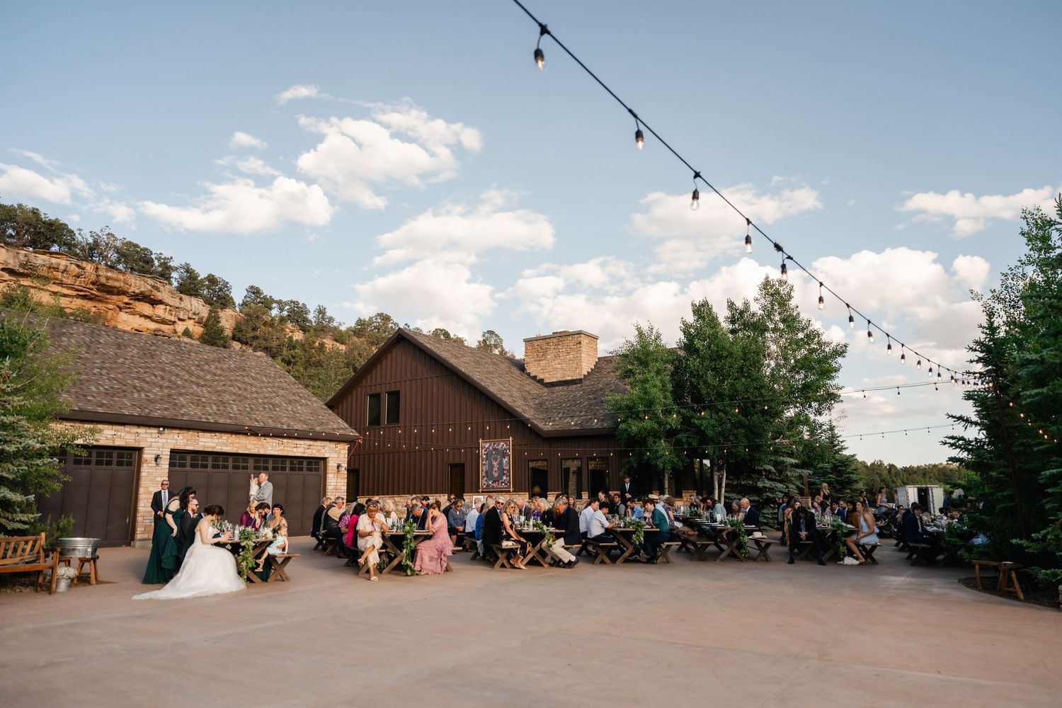 A panoramic view of an outdoor wedding reception venue with string lights and guests gathered on a patio.