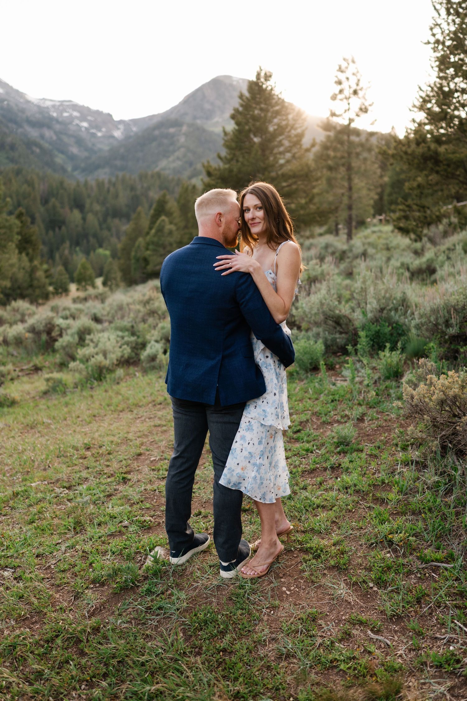 Couple embracing at golden hour in a mountain meadow, woman in floral dress and man in navy suit, pine trees behind.