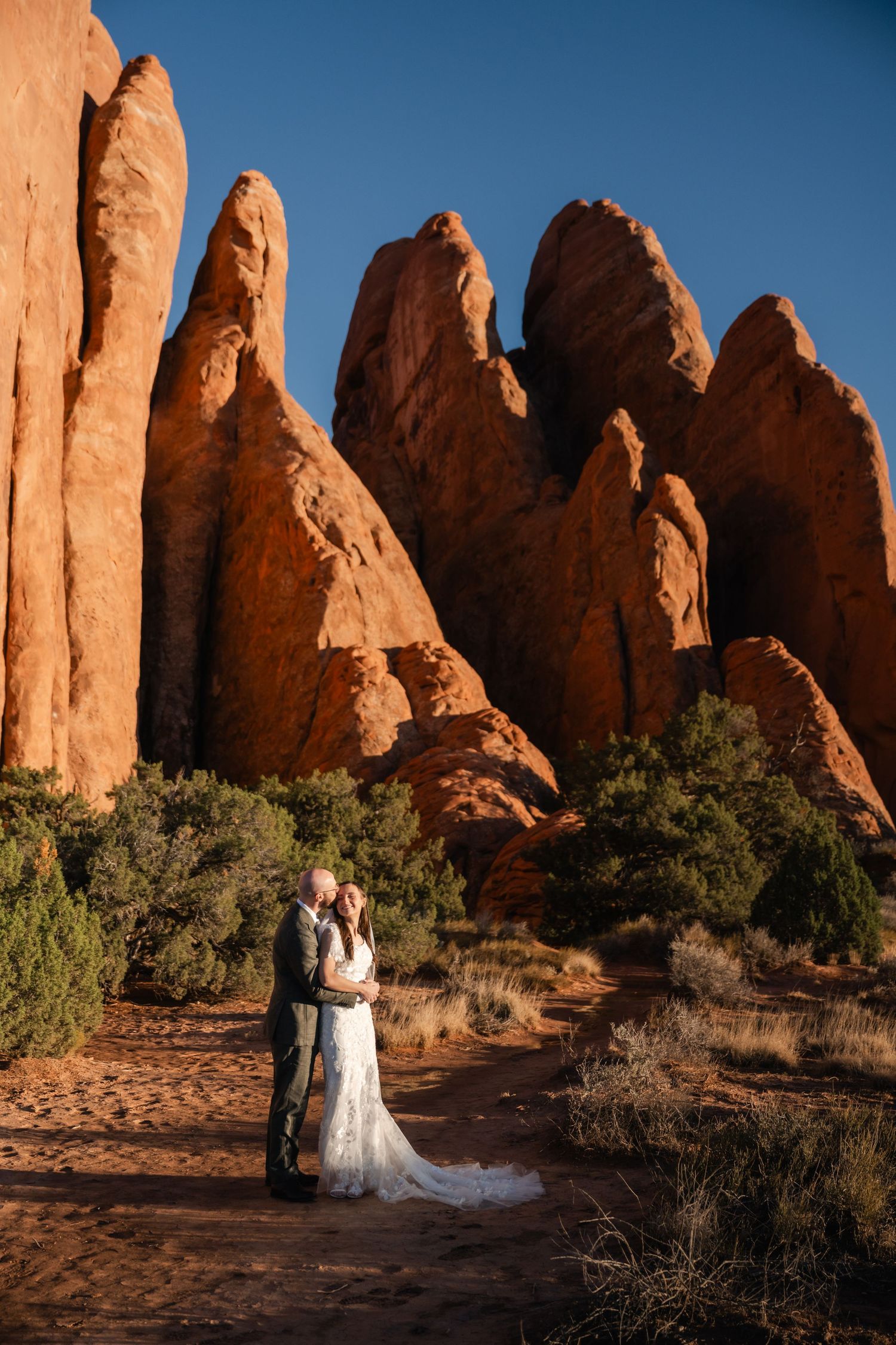 A couple in wedding attire stands against towering red rock formations at Arches National Park in Utah.