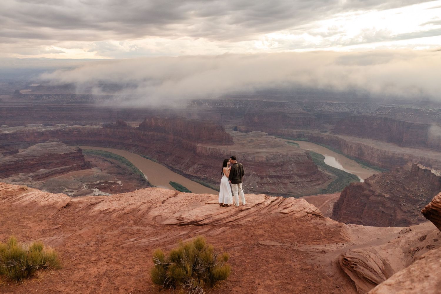 Two people stand at Dead Horse Point State Park overlooking the dramatic Colorado River bend during a cloudy sunset.