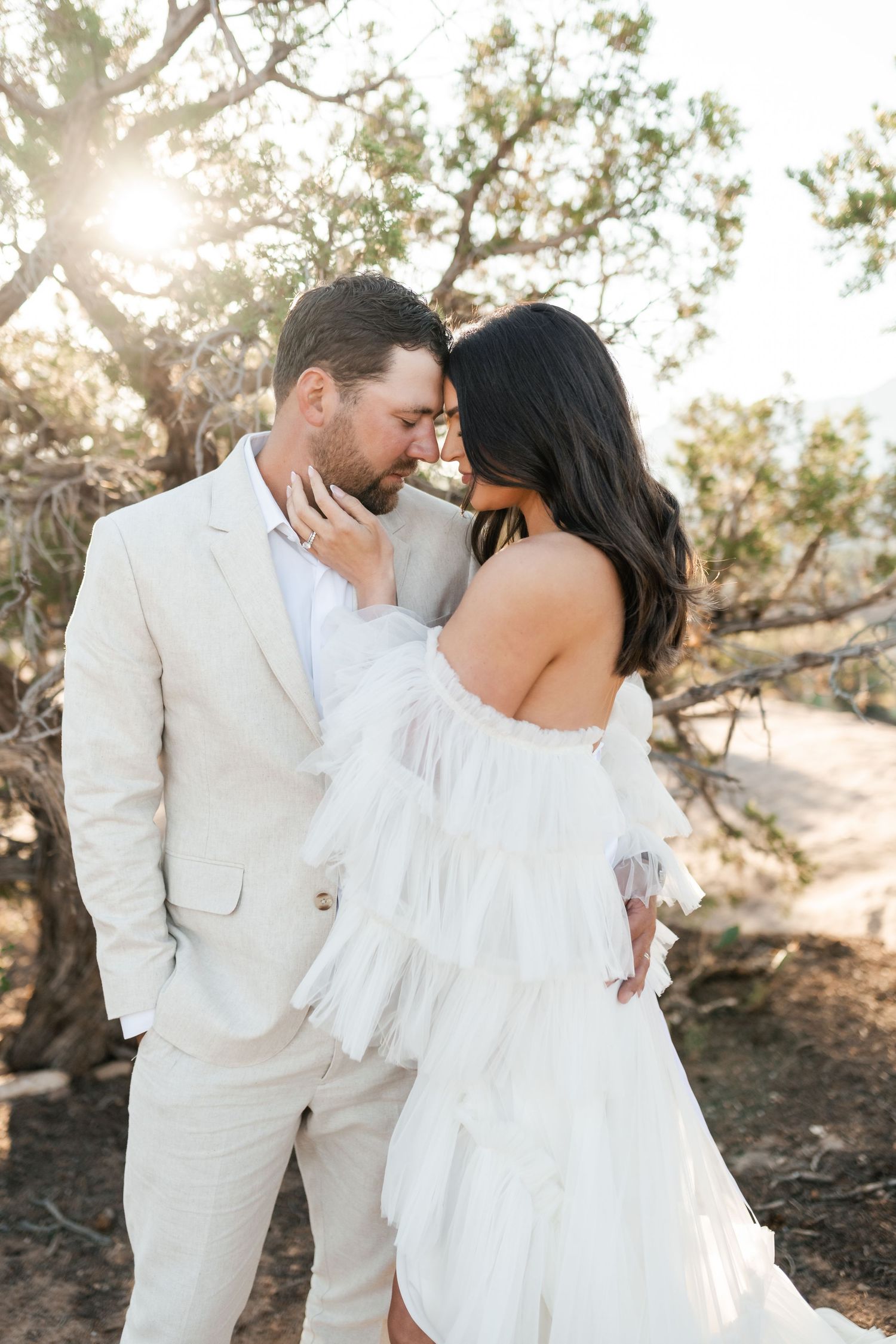 Zion destination photographer, A romantic couple in white attire embracing among desert trees at sunset.
