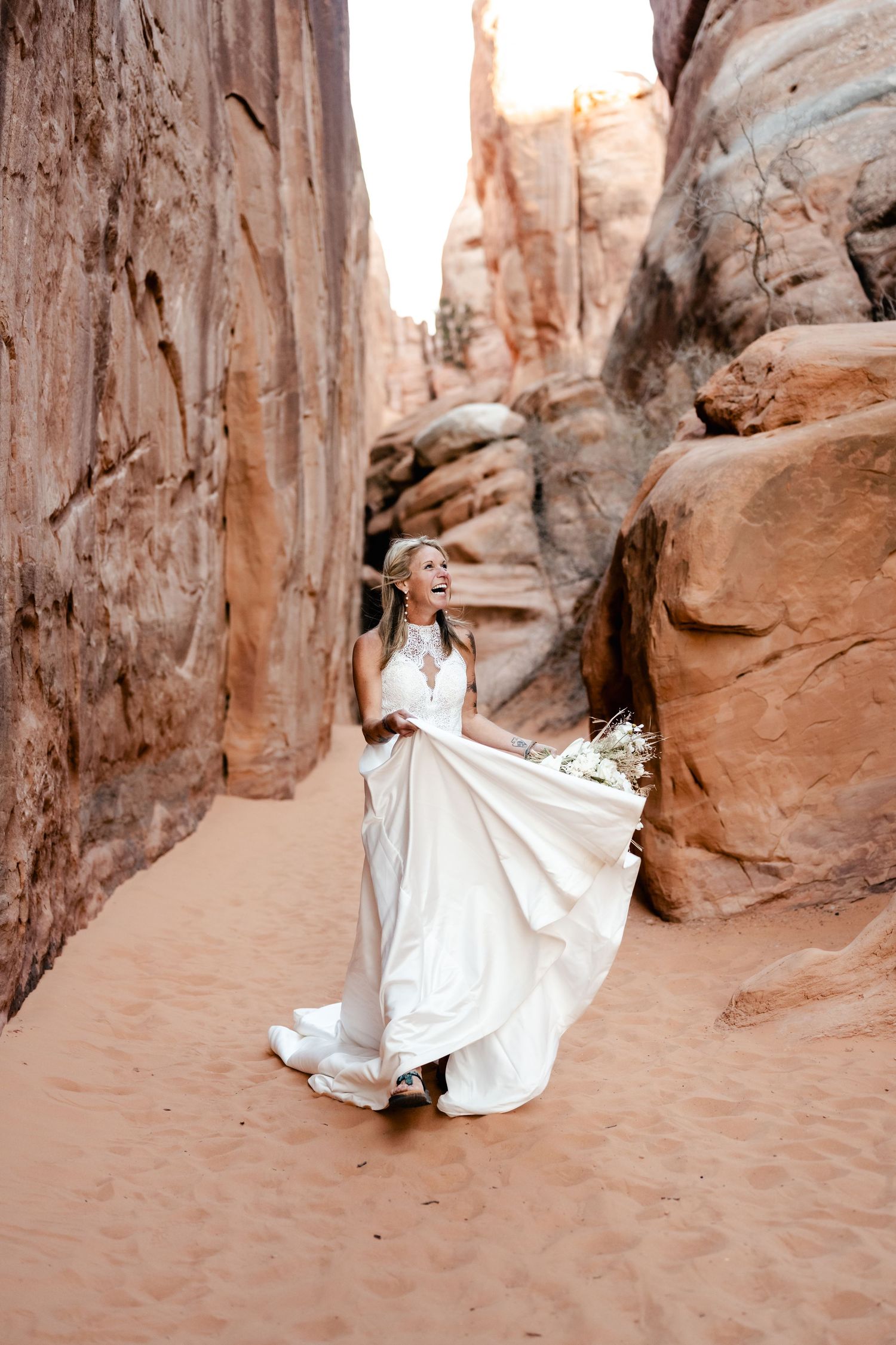 A bride in a flowing white dress twirls and dances among towering desert canyon walls.