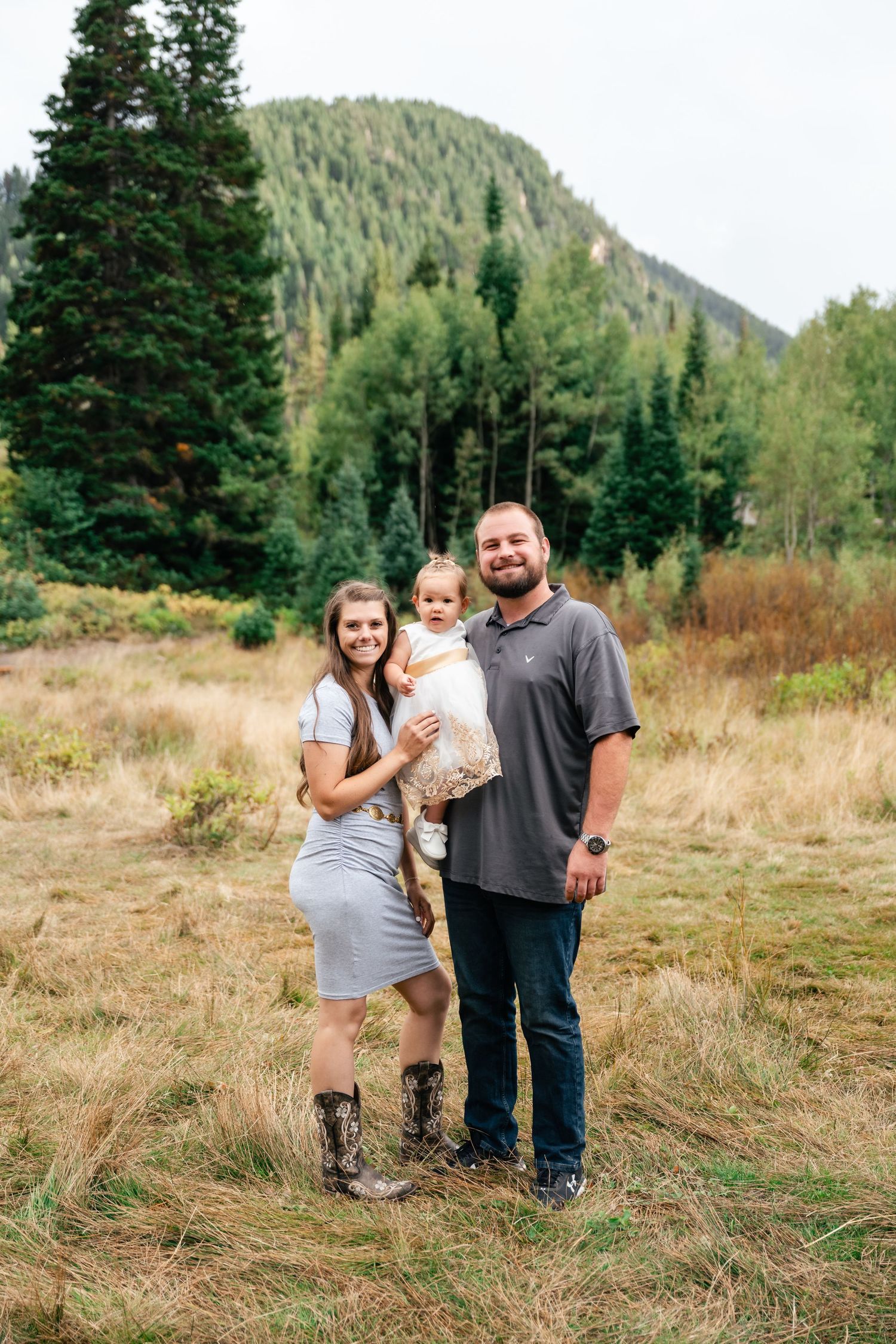 Family of three poses together in mountain meadow with evergreen forest and peaks in background.