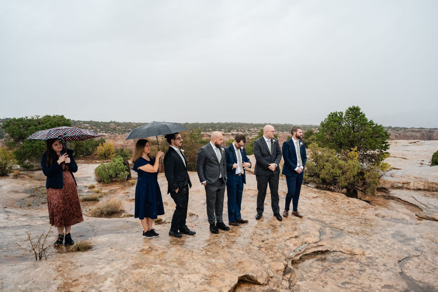 Wedding party standing on rocky terrain under cloudy skies in formal attire.