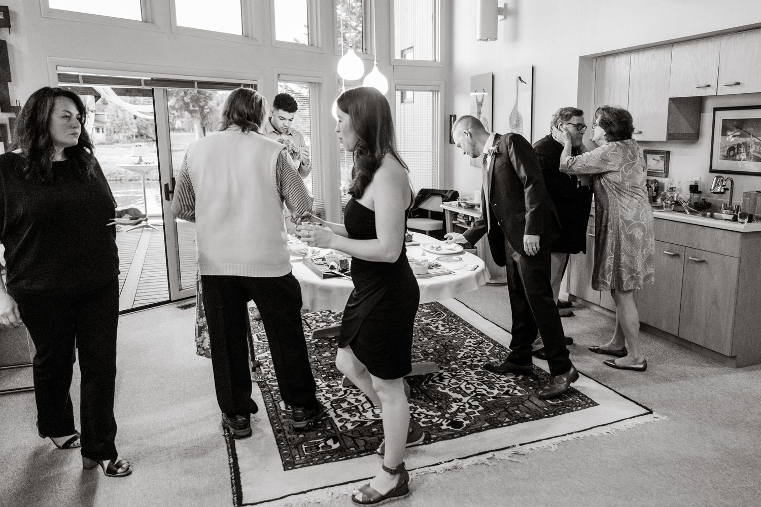 Black and white photo of people mingling at an indoor event near large windows.