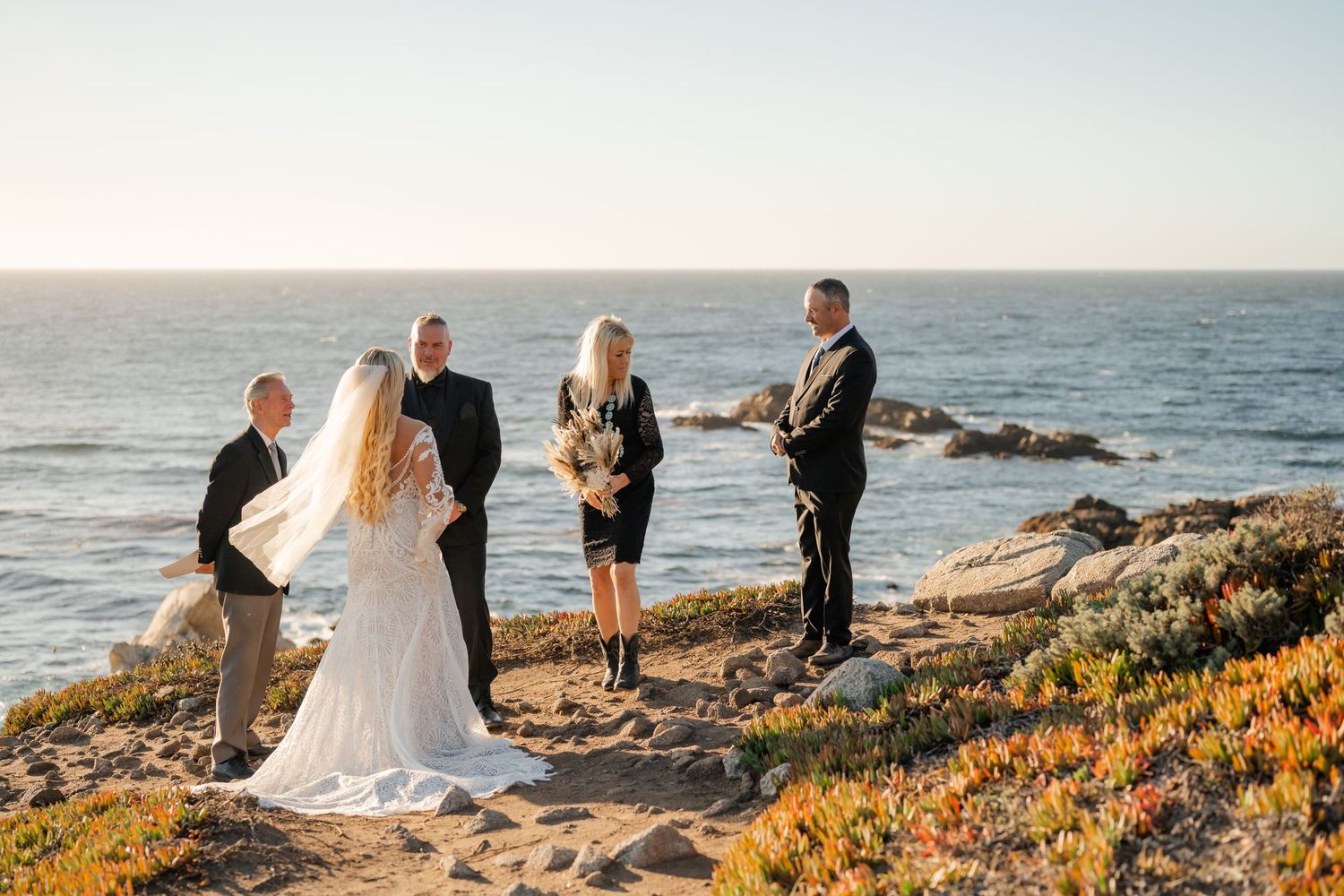 A small wedding party gathers on a coastal bluff with waves crashing against rocks in the background.