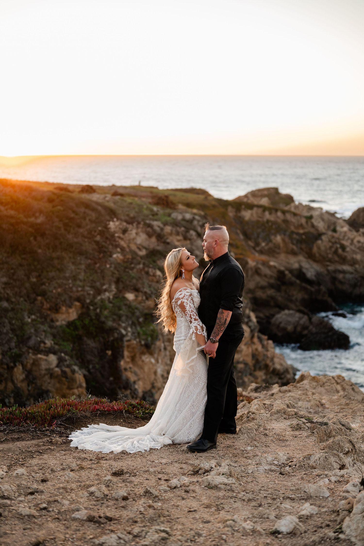 A romantic coastal wedding portrait at golden hour with rocky cliffs and ocean backdrop.