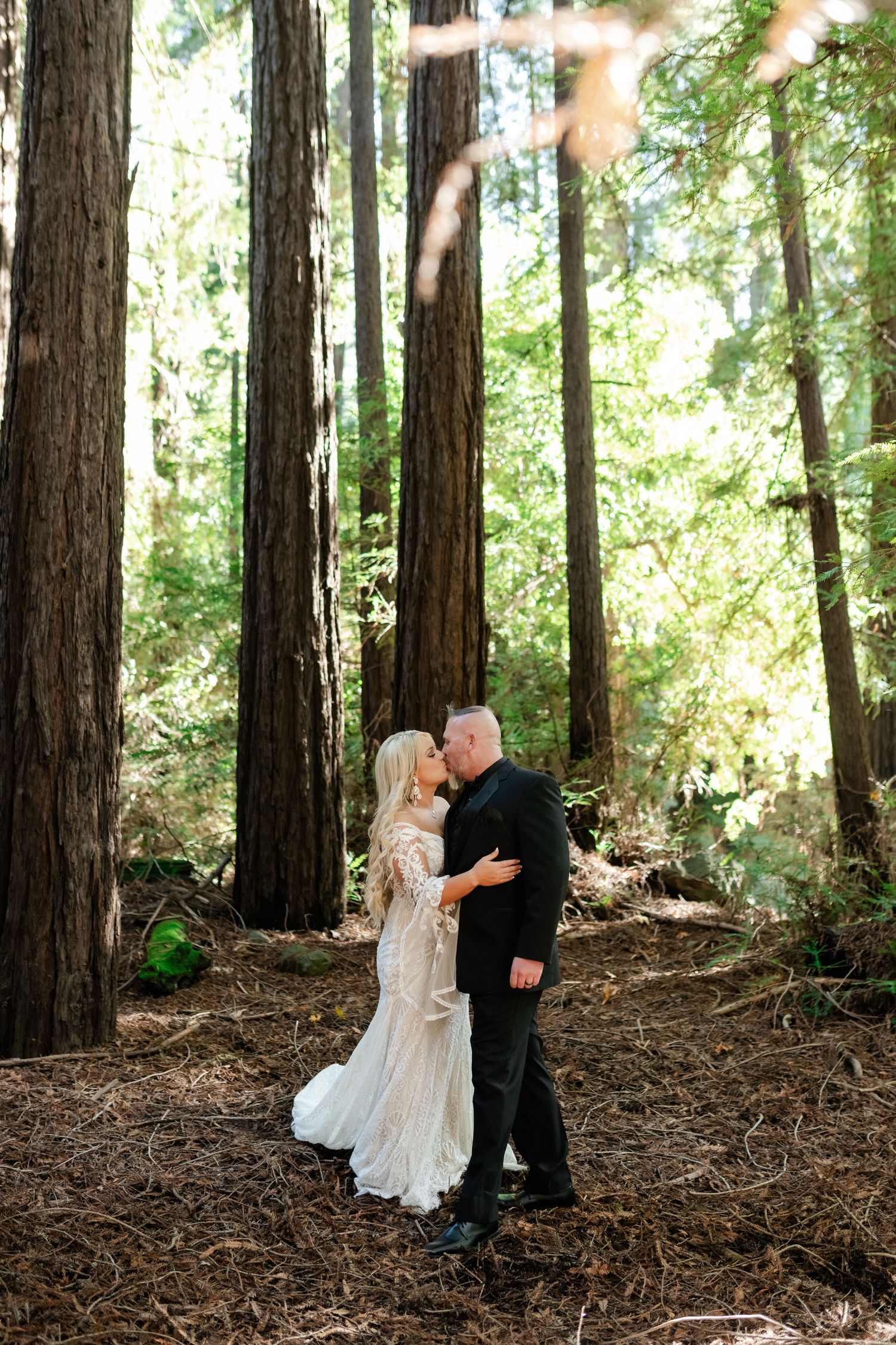 A romantic wedding moment captured in a sunlit redwood forest with tall majestic trees.