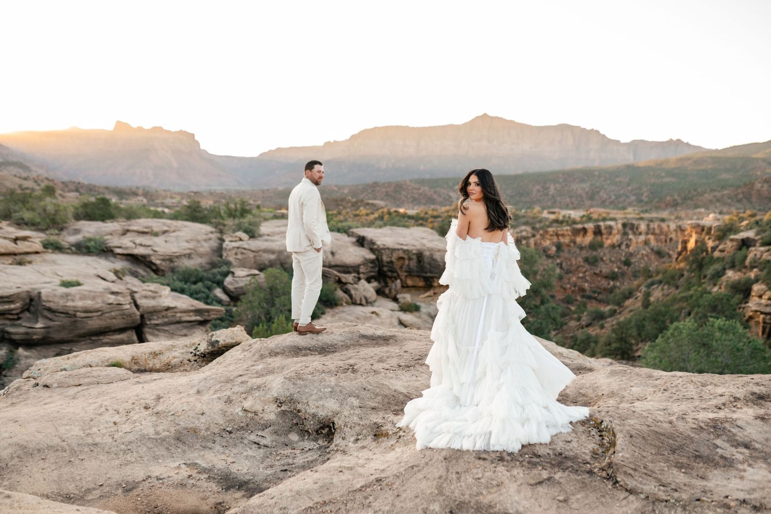 A couple in wedding attire stands on red rock cliffs overlooking a scenic desert canyon landscape at sunset. Zion elopement photographer.