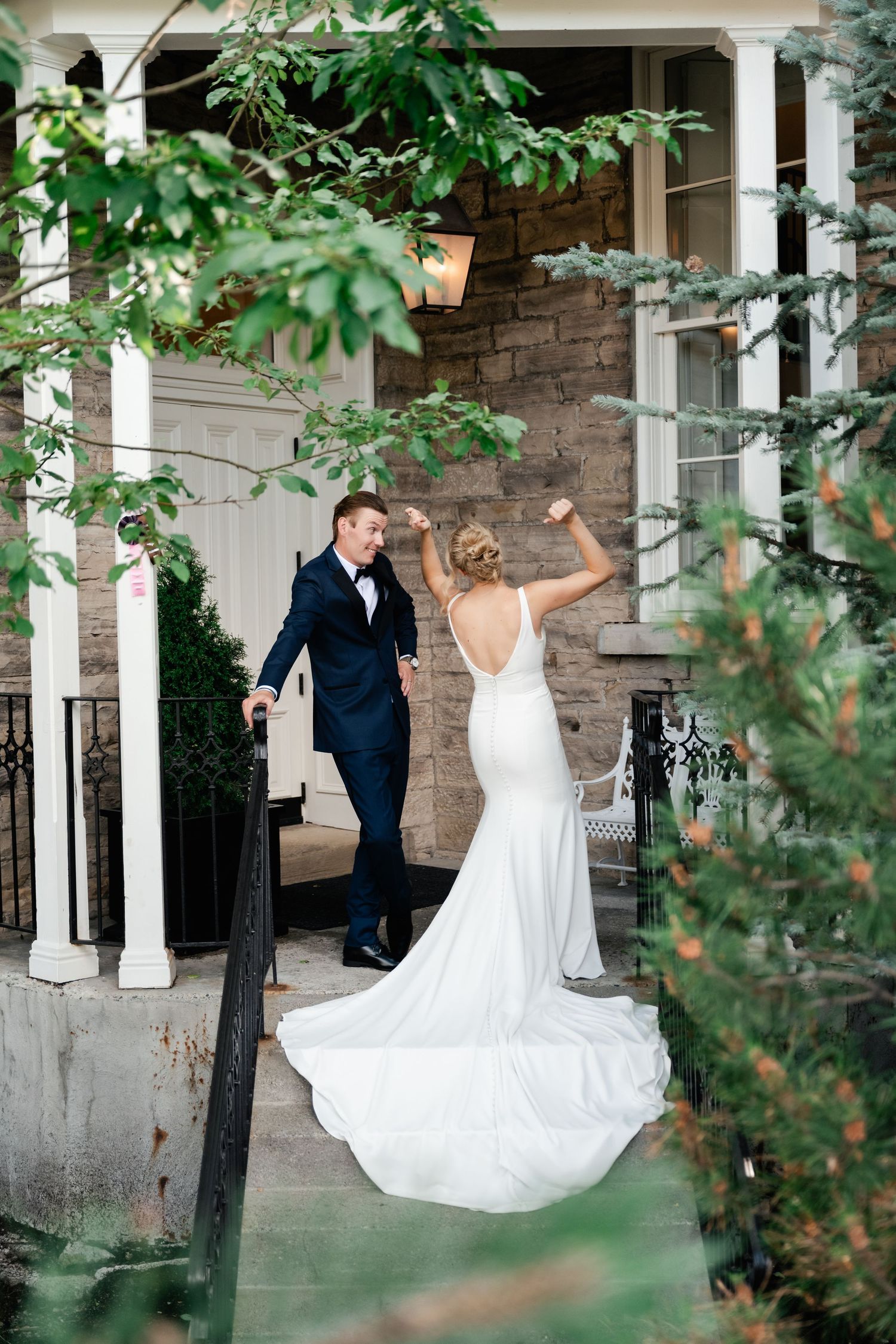 Playful wedding couple dancing under a green-covered porch with stone columns.