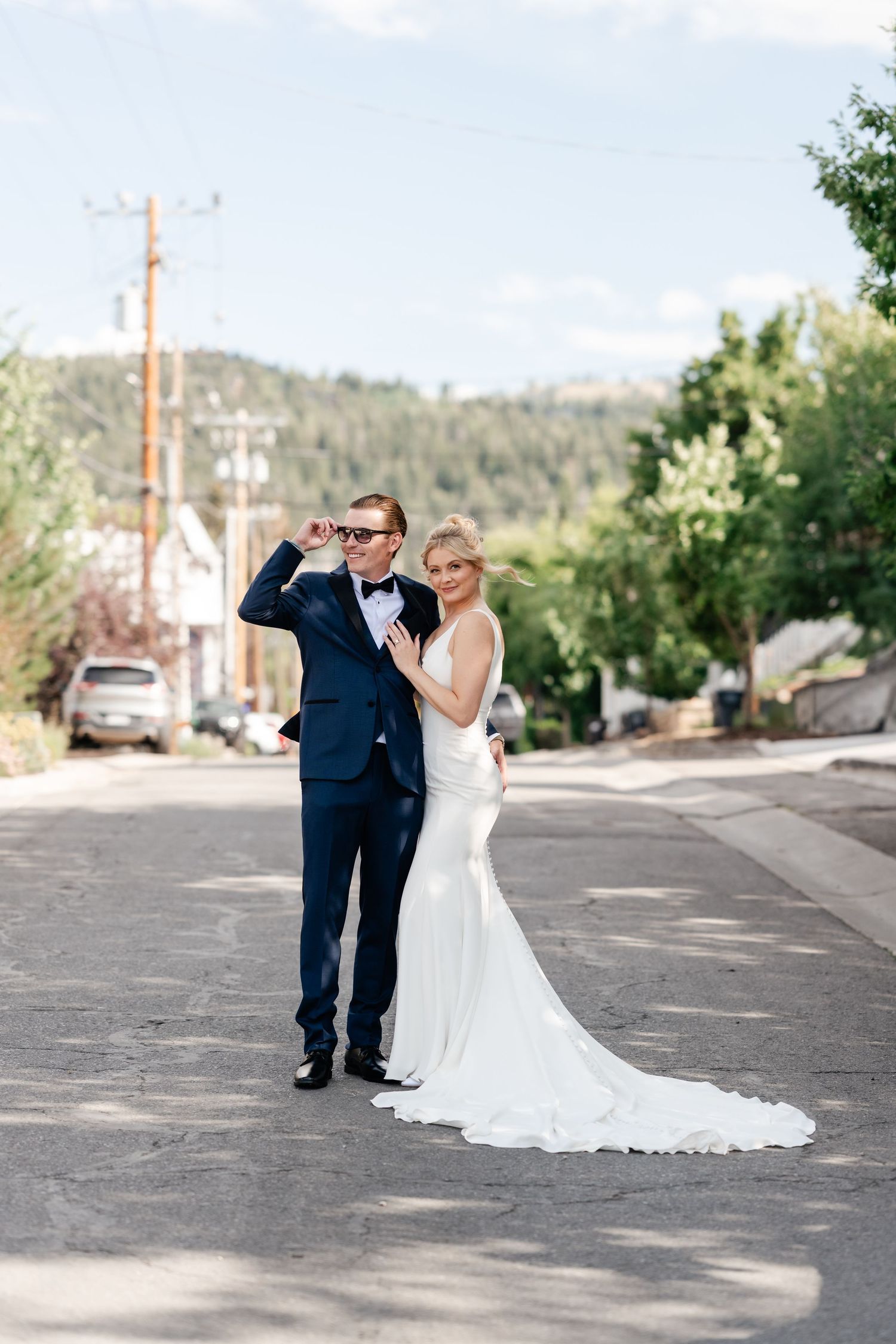 Wedding couple poses playfully on sunny small town street with mountains in background.