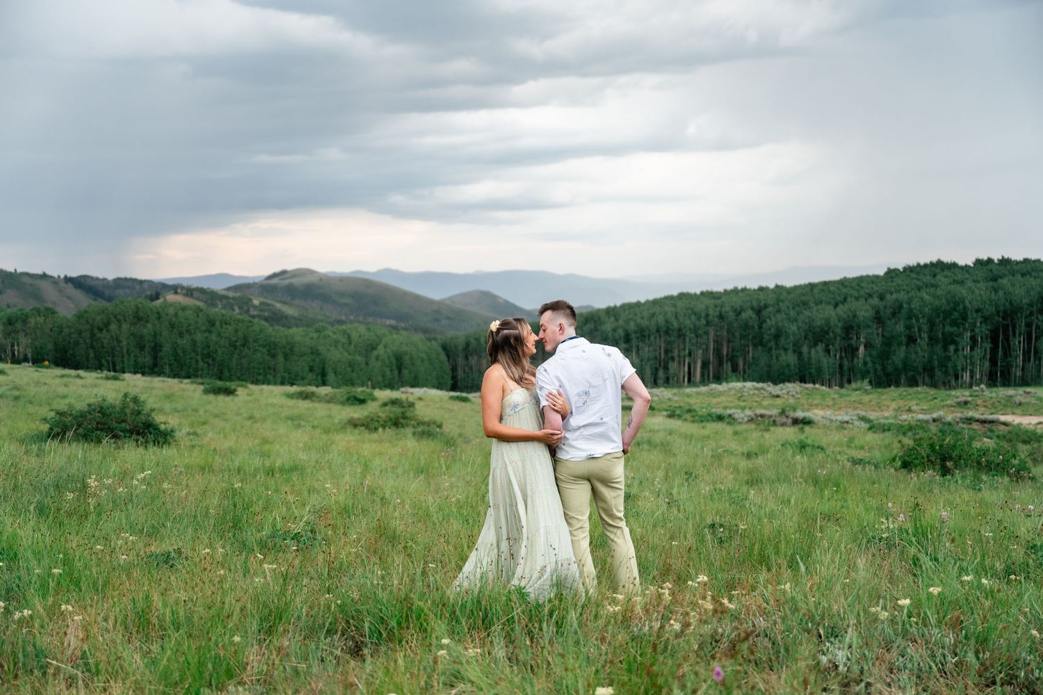 A couple shares a romantic moment in a scenic mountain meadow with rolling green hills and cloudy skies in the background.