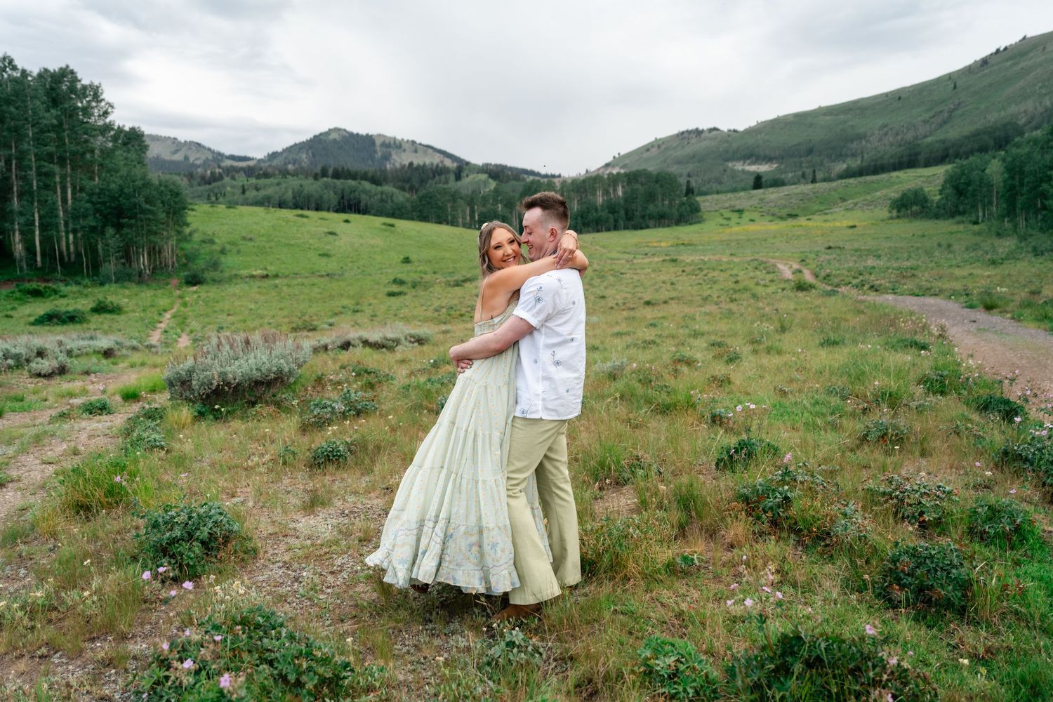 Couple embraces in a scenic mountain meadow surrounded by evergreen forests and rolling hills.