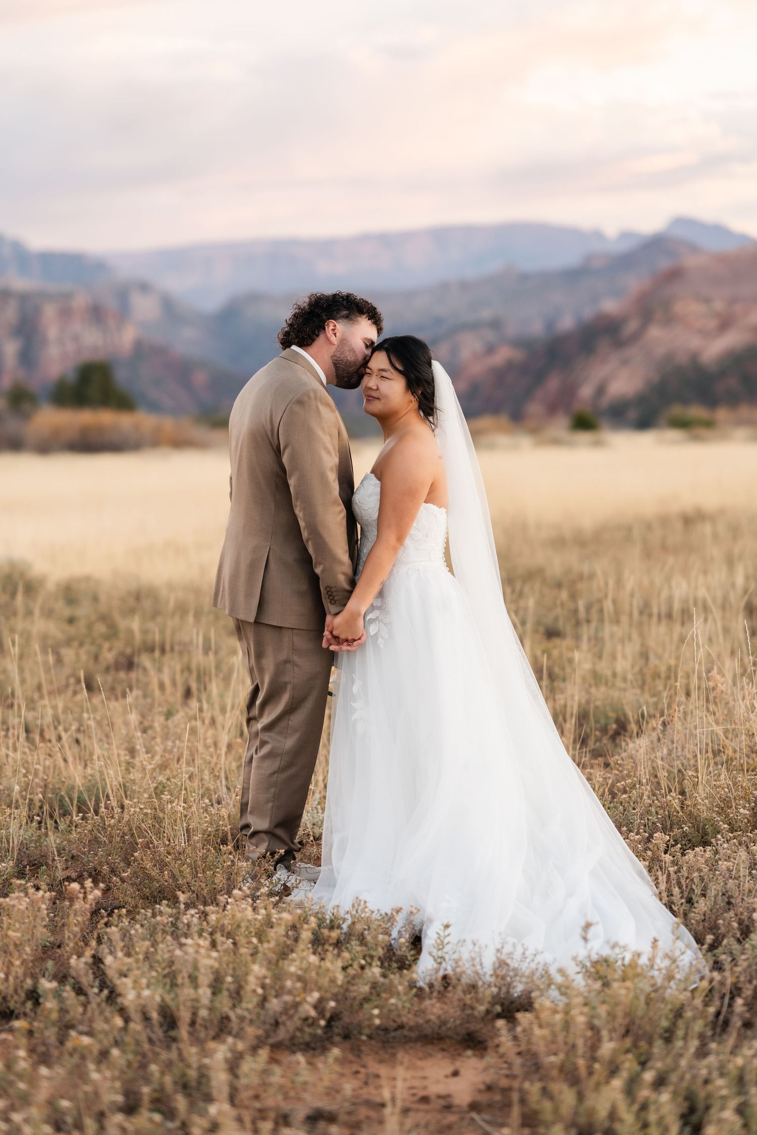 A romantic wedding photoshoot in a golden field with mountains in the background at dusk.