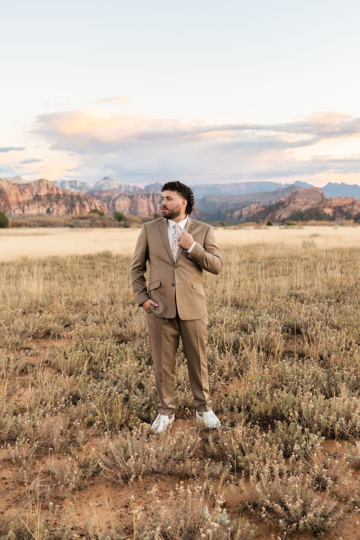A couple in wedding attire poses playfully in a desert field with mountains on the horizon.