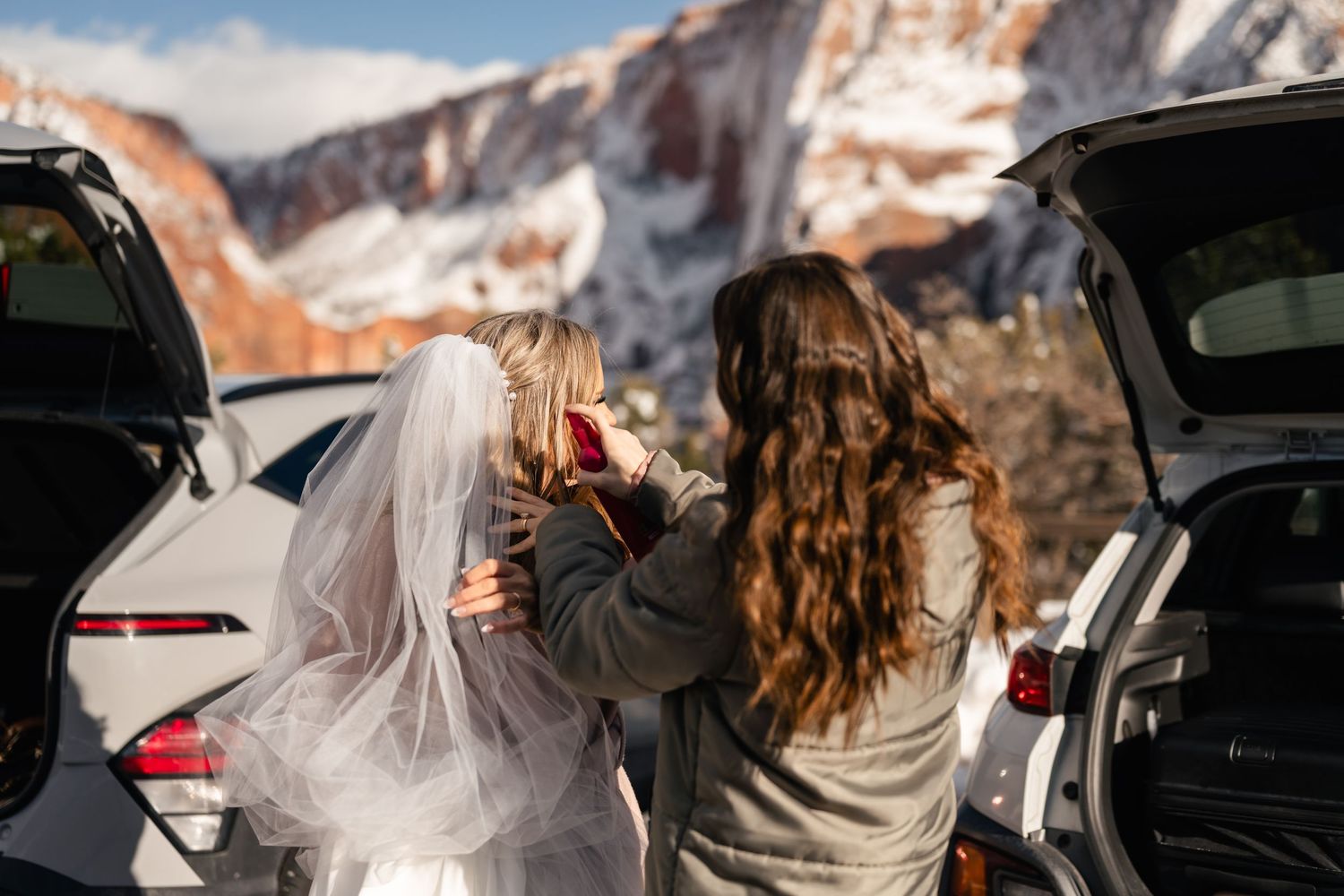 Two people prepare for a wedding ceremony near vehicles with snowy mountains in background.