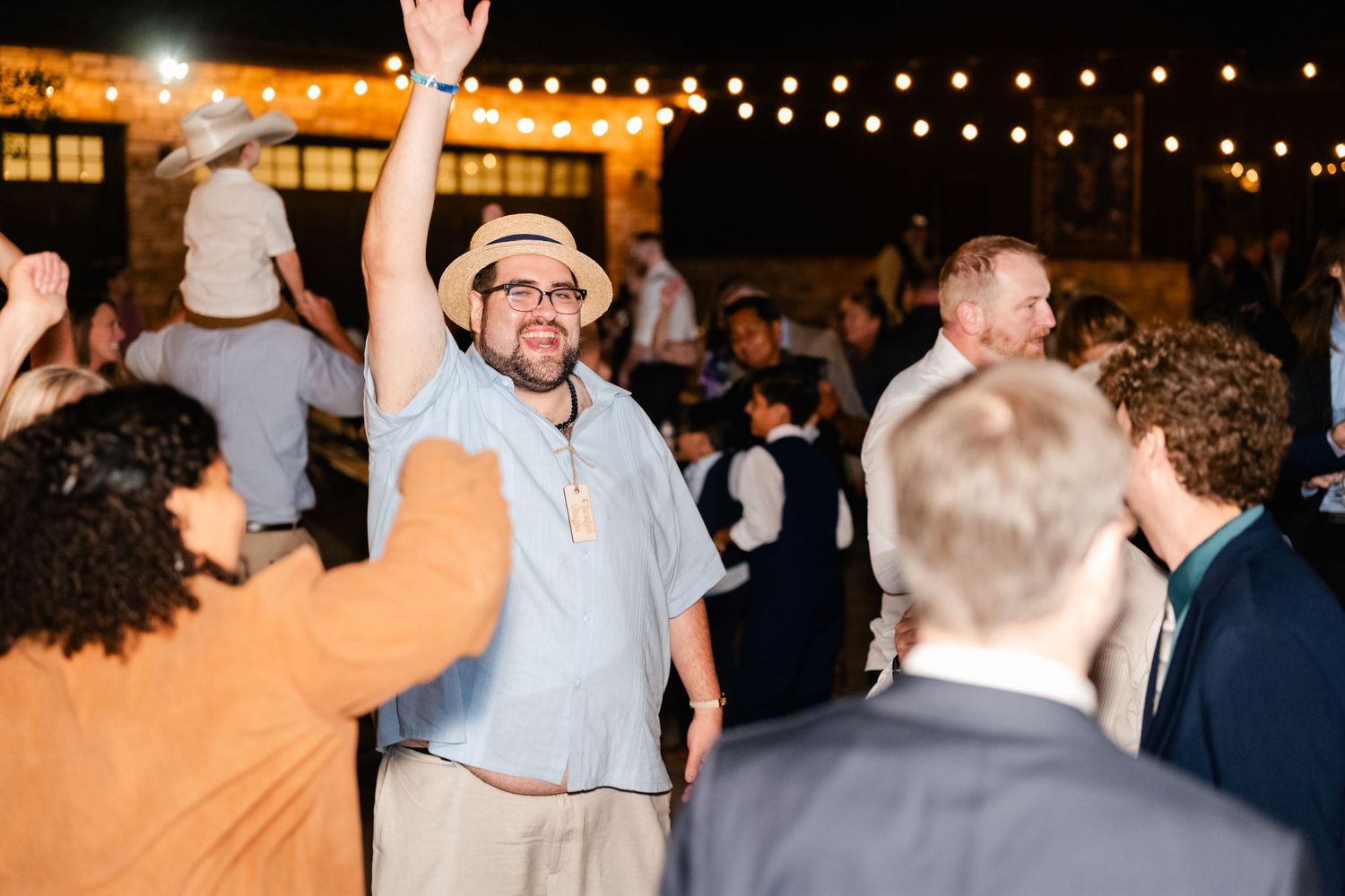 Crowd of wedding guests dancing and raising their hands under glowing string lights at an evening reception celebration.
