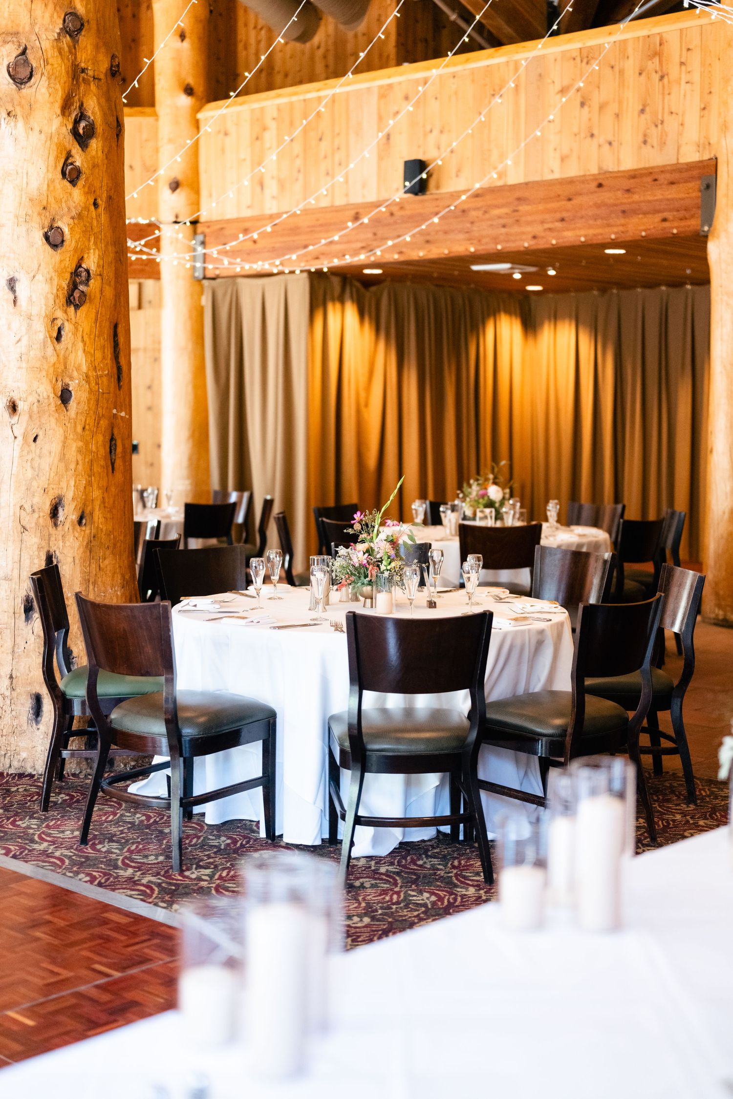 Rustic wedding reception venue with wooden beams, white tablecloths and dark chairs under warm lighting.