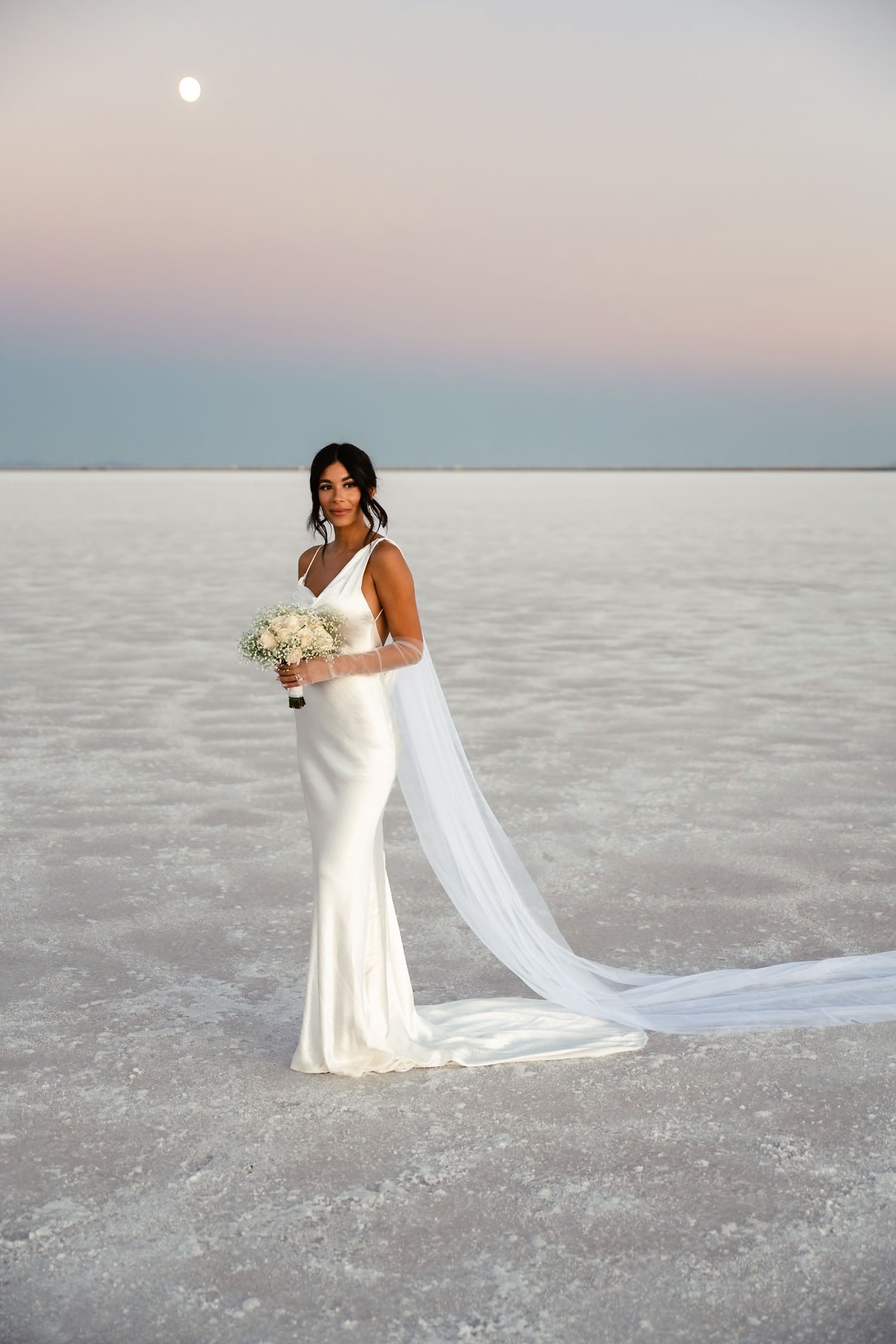 A bride poses in an elegant white wedding dress with flowing veil on salt flats under pastel skies.