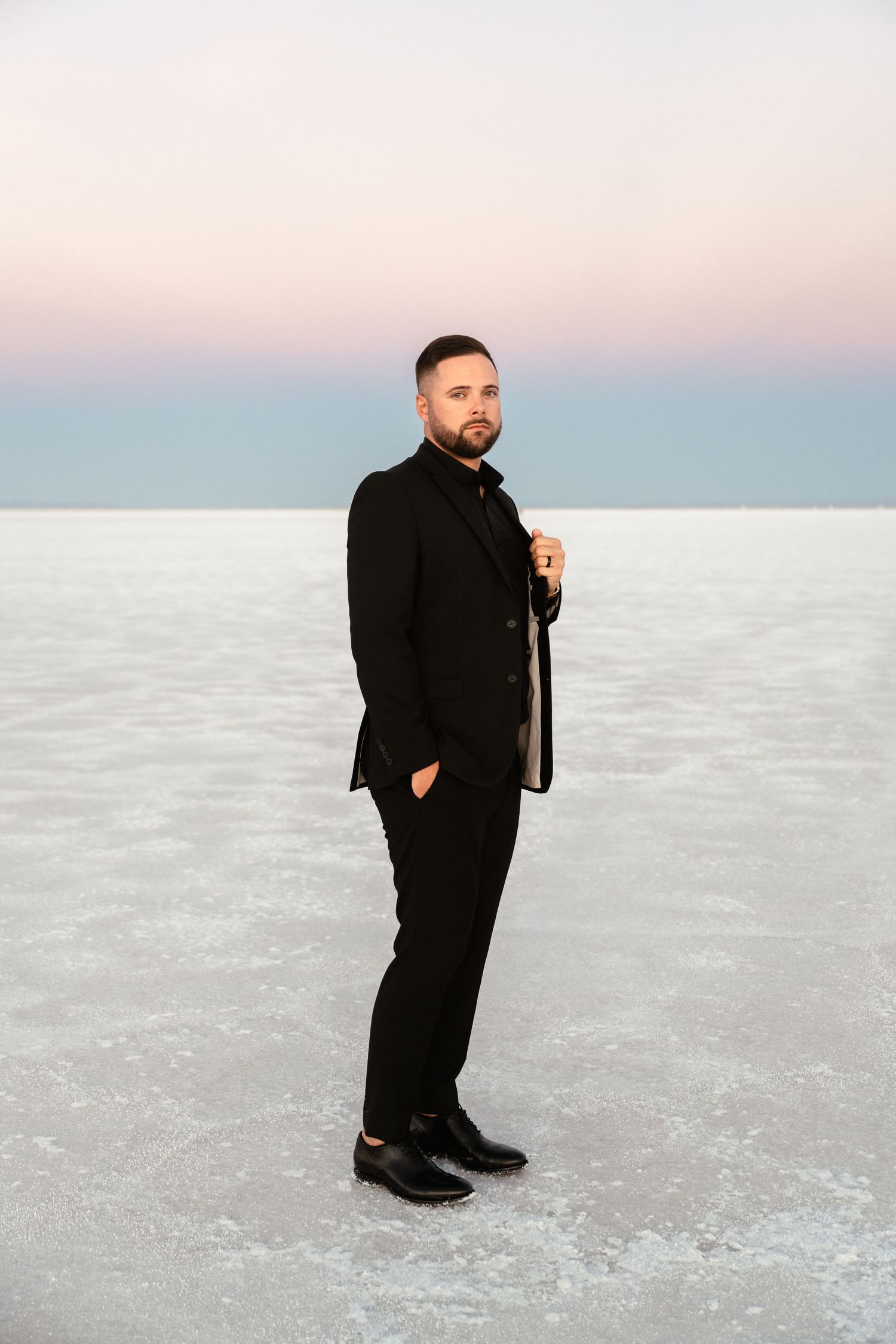 A person in black formal attire poses on a white salt flat landscape at sunset.