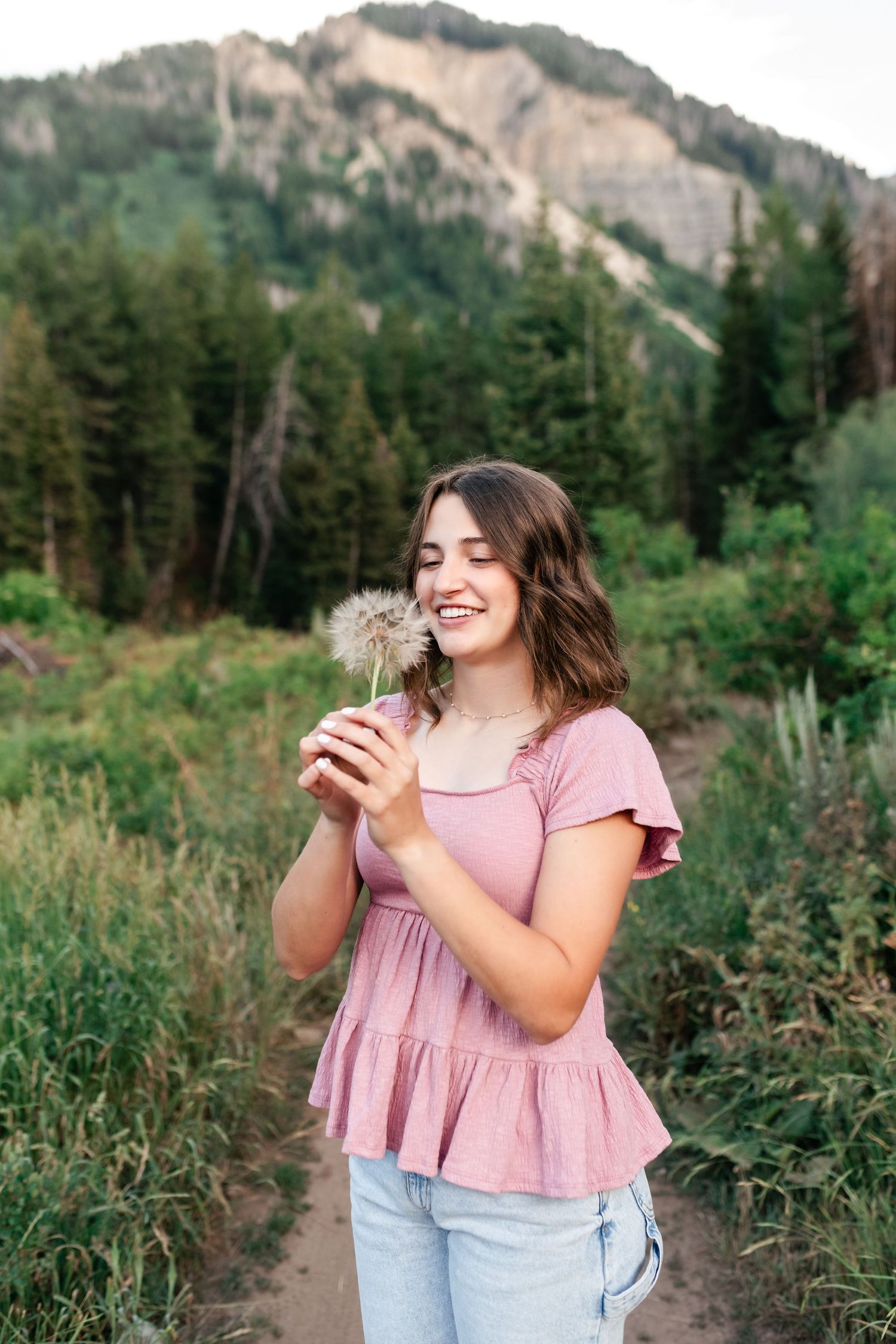 Someone wearing a pink top holds and blows on a dandelion in a mountain meadow.