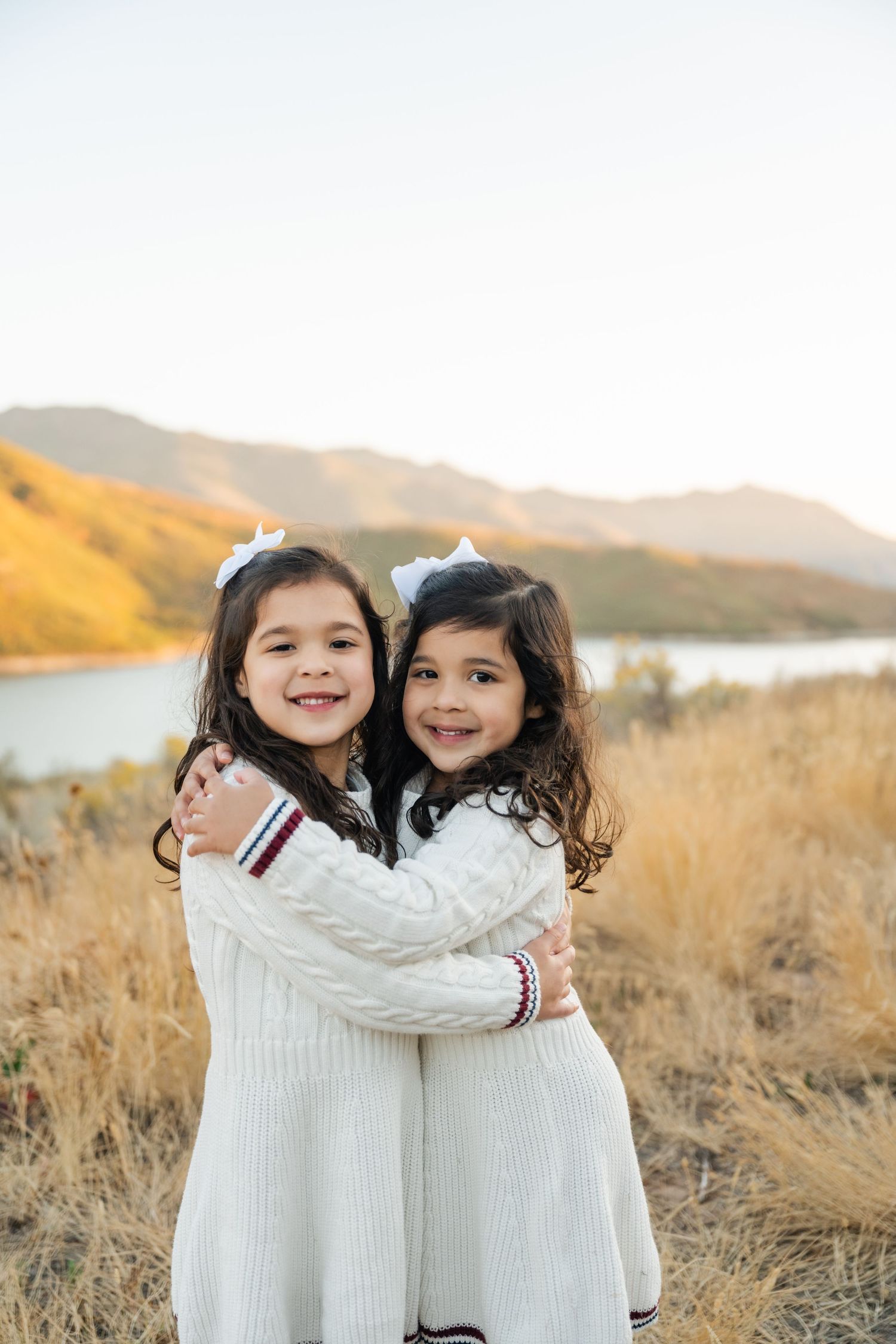 Two children in matching white sweaters embrace and smile while standing in a golden field at sunset.