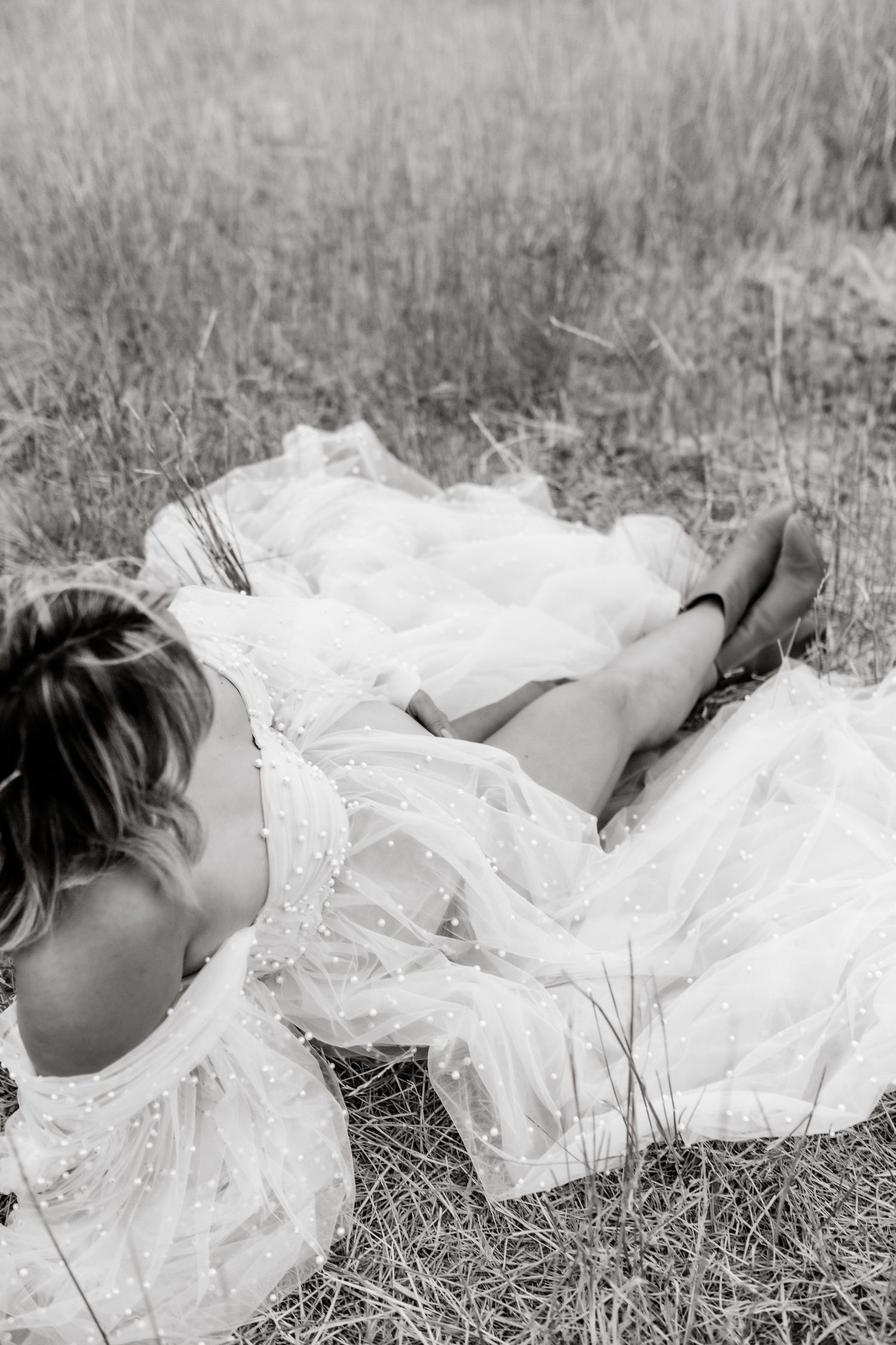 A black and white artistic photograph shows someone lying in tall grass wearing a flowing white lace dress.