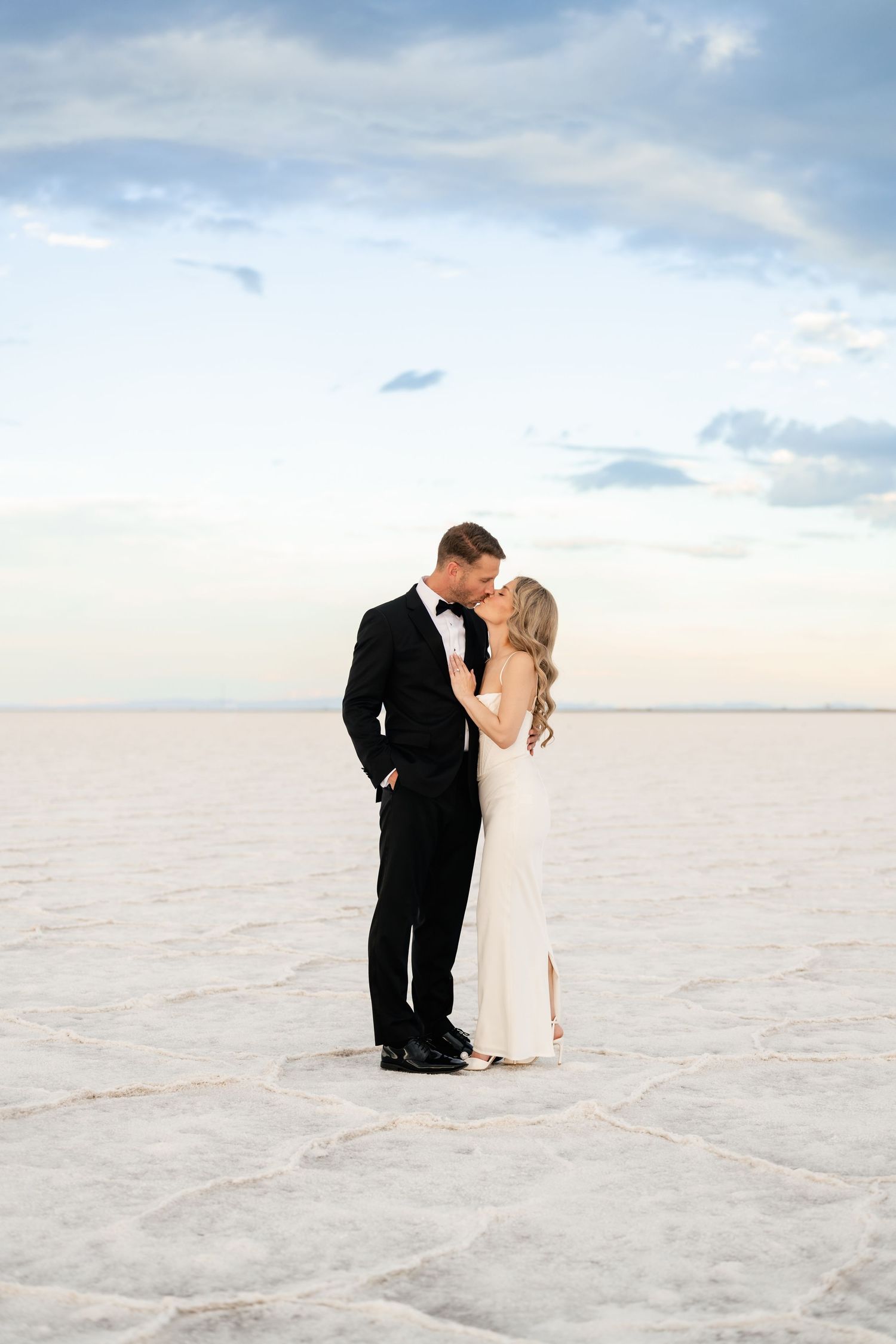 Salt Flats Elopement Photographer. Newlyweds in formal attire embrace on a stark white salt flat landscape under cloudy skies.