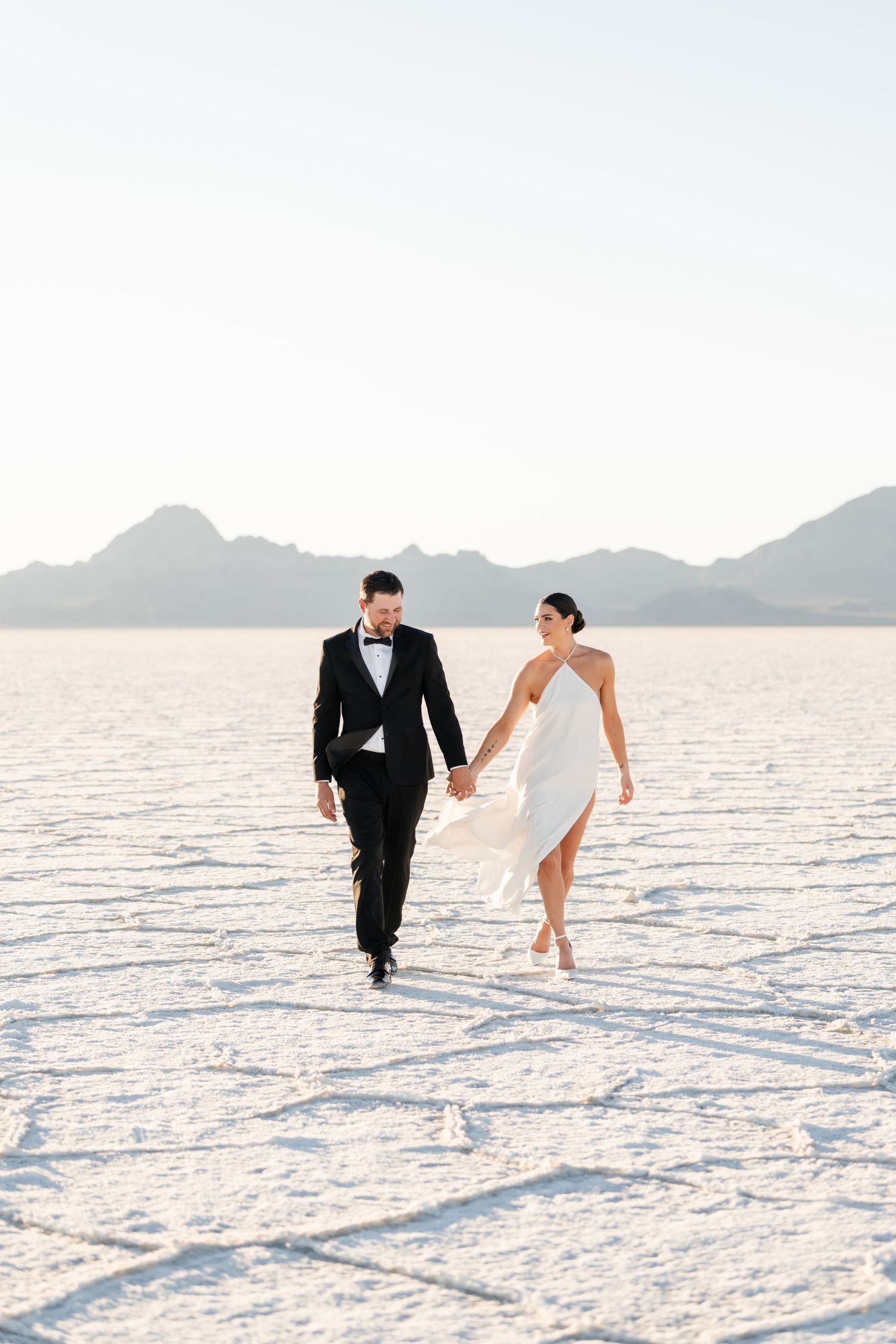 Salt Flats Photographer. A couple runs playfully across white salt flats under a bright blue sky with mountains in the distance.