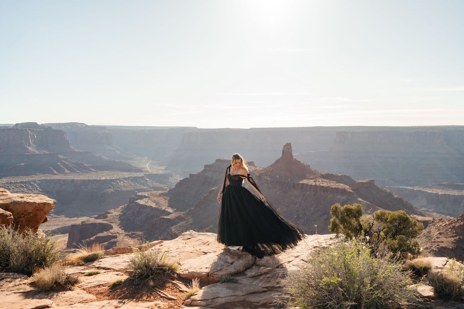 A person in a flowing black dress stands on a rocky outcrop overlooking the dramatic canyons and mesas of Dead Horse Point.