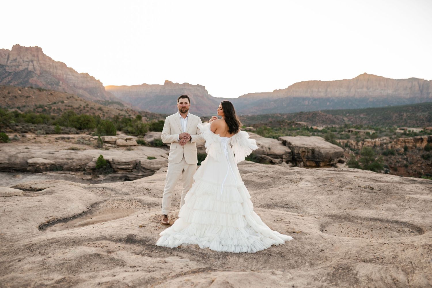 Couple in wedding attire poses on rocky desert terrain with scenic mountain landscape at sunset.