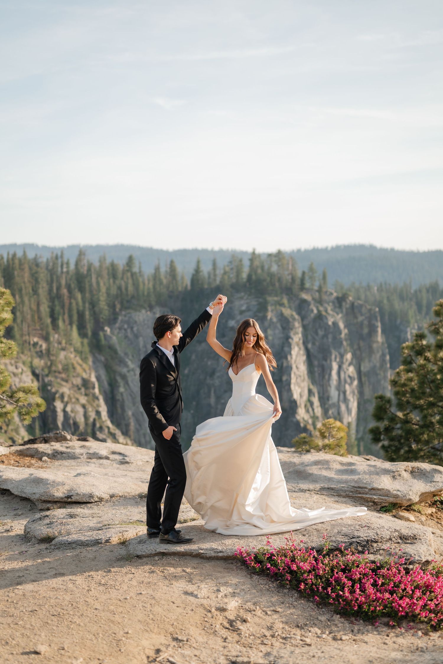Bride and groom dancing on rocky cliff at Yosemite with mountain forest backdrop and pink wildflowers.