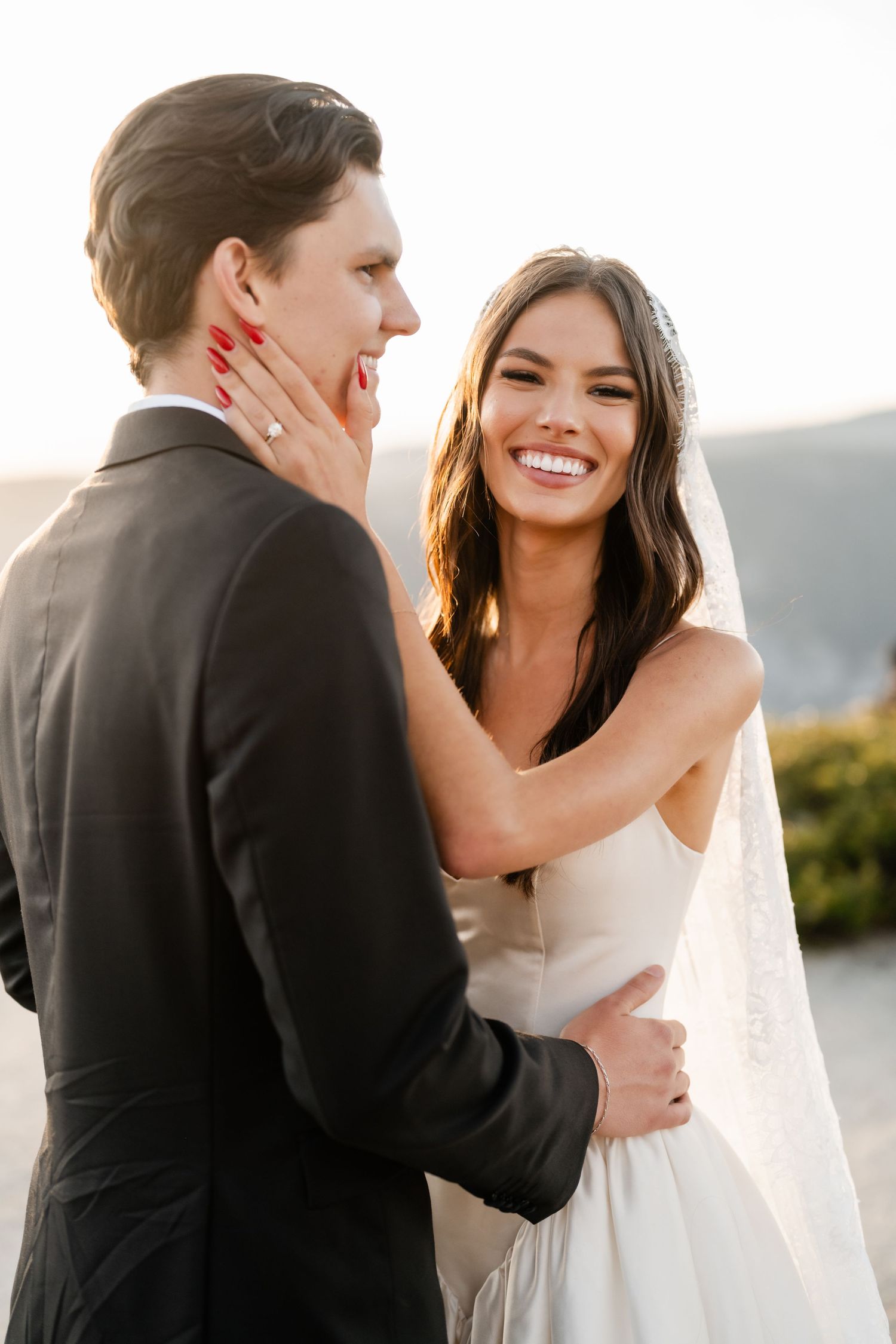 Smiling bride in white gown and veil tenderly touches groom's face during golden hour outdoor wedding ceremony.