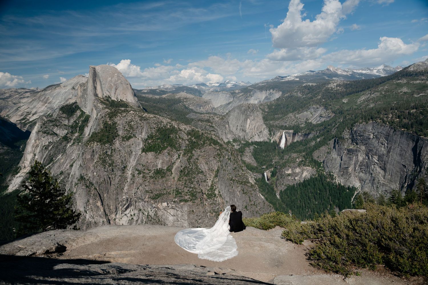 Couple in wedding attire sitting on rocky cliff at Yosemite with Half Dome and sweeping valley views behind them.